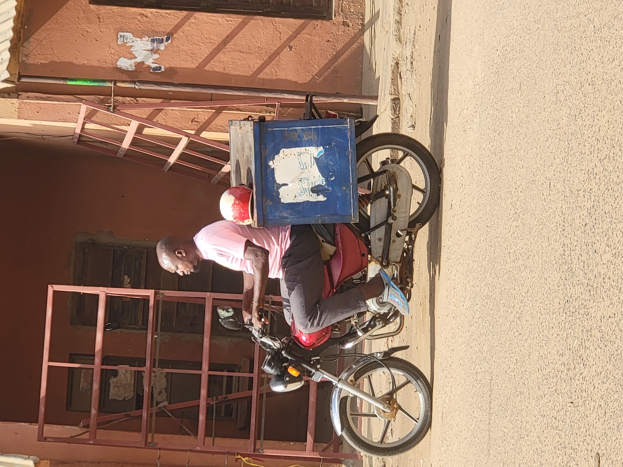 The image depicts a man riding a motorcycle in Maiburiji, Nigeria. The man is wearing a pink shirt, gray pants, and blue sneakers. He also wears a red and white helmet. A blue delivery box is mounted on the back of the motorcycle. The buildings in the background are made of red brick and have windows with metal bars. The street is paved with concrete.