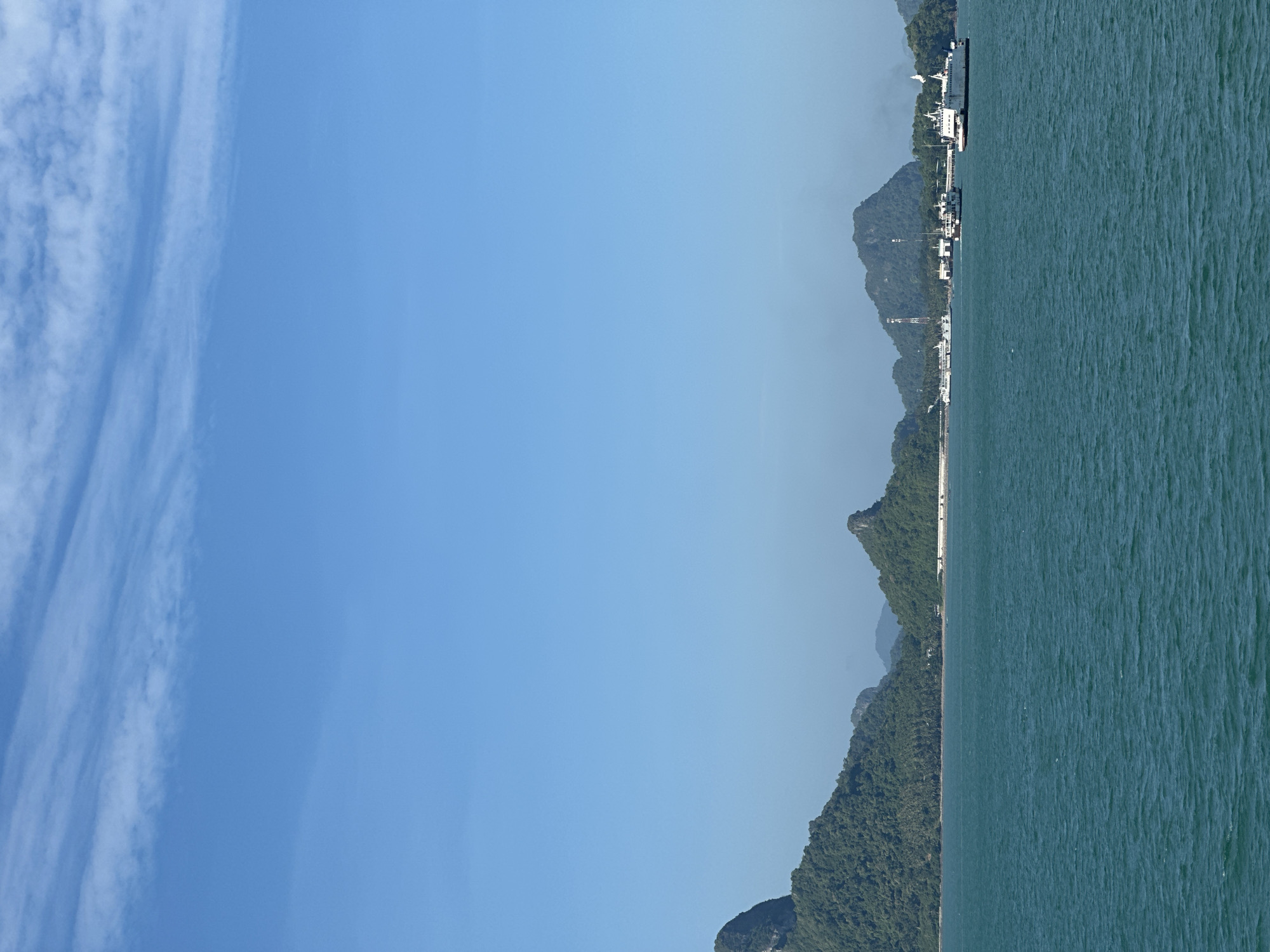 The image shows a wide view of the sea with islands in the distance. The water is a vibrant teal color and appears somewhat choppy. In the background, a chain of lush green islands with steep, rocky peaks stretches across the horizon. Some of the islands are covered in dense vegetation, while others appear more barren and rocky. A small town or village is visible nestled along the coastline of one of the islands, with buildings, a pier, and a tall communication tower. Several ferries are present in the water, with one large white ferry prominently docked or moving near the shore. The sky is a clear, bright blue with a few wispy white clouds. The scene suggests a coastal or island location, possibly a port or ferry terminal. This scene is typical of the coastline of Don Sak, Thailand, a region known for its picturesque islands and ferry services connecting to islands like Koh Samui and Koh Phangan.
