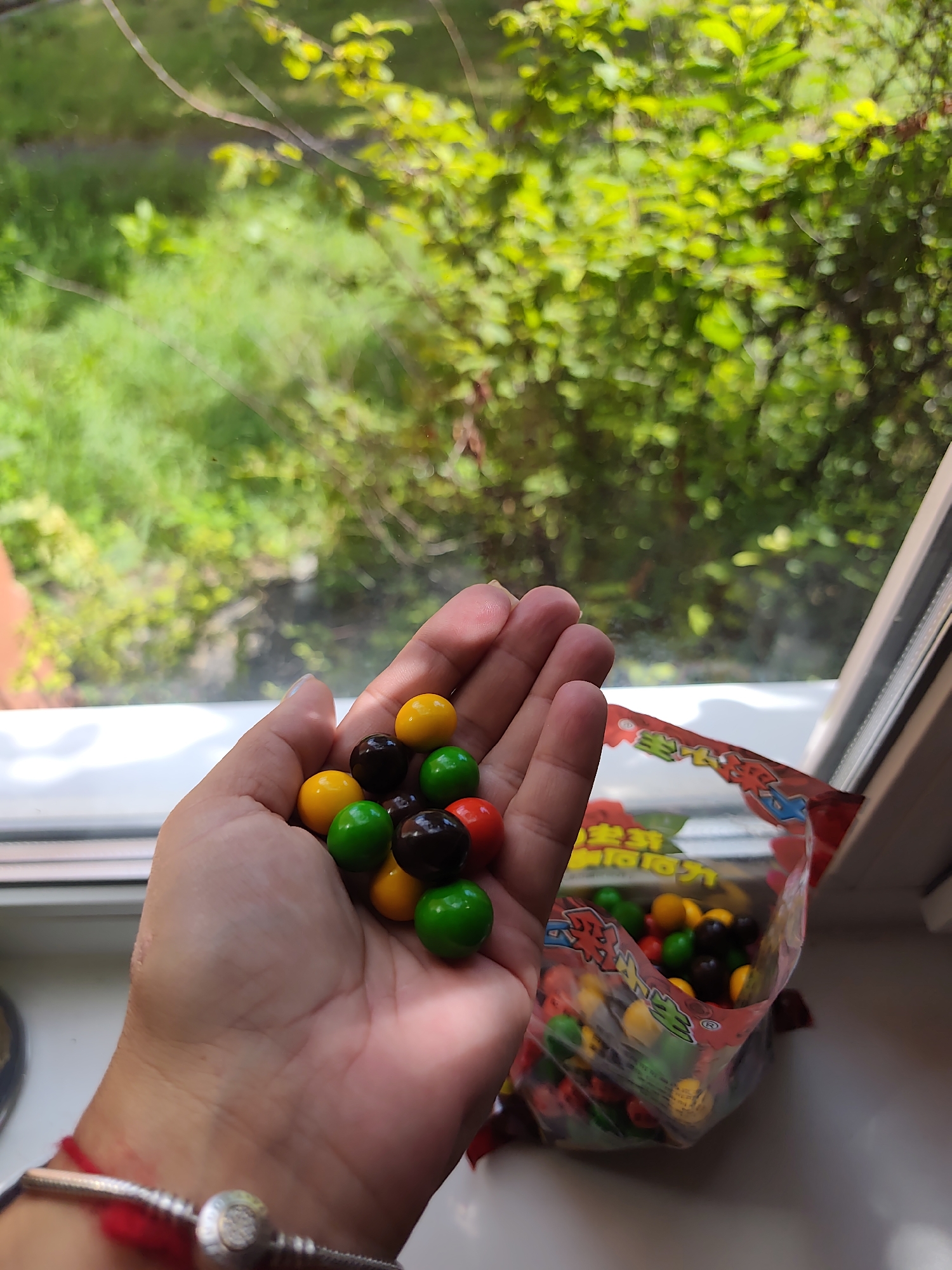 A hand holds a collection of brightly colored candies, predominantly yellow, green, and brown, with a few red ones visible. The hand is positioned in the foreground, slightly to the left of the center of the frame. Behind the hand, to the right, a bag of similar candies is partially visible, its red and yellow packaging suggesting it is a snack item. The background reveals a window with a view of lush green foliage and branches, indicating an outdoor setting. The windowsill where the hand and candy bag rest appears to be made of a white material, possibly plastic. A thin red string is tied around the wrist of the hand, accompanied by a silver charm bracelet. The lighting suggests it is daytime, with sunlight filtering in from the window. The scene is devoid of any explicit indications of the city of Dzerzhinsk, Russia.