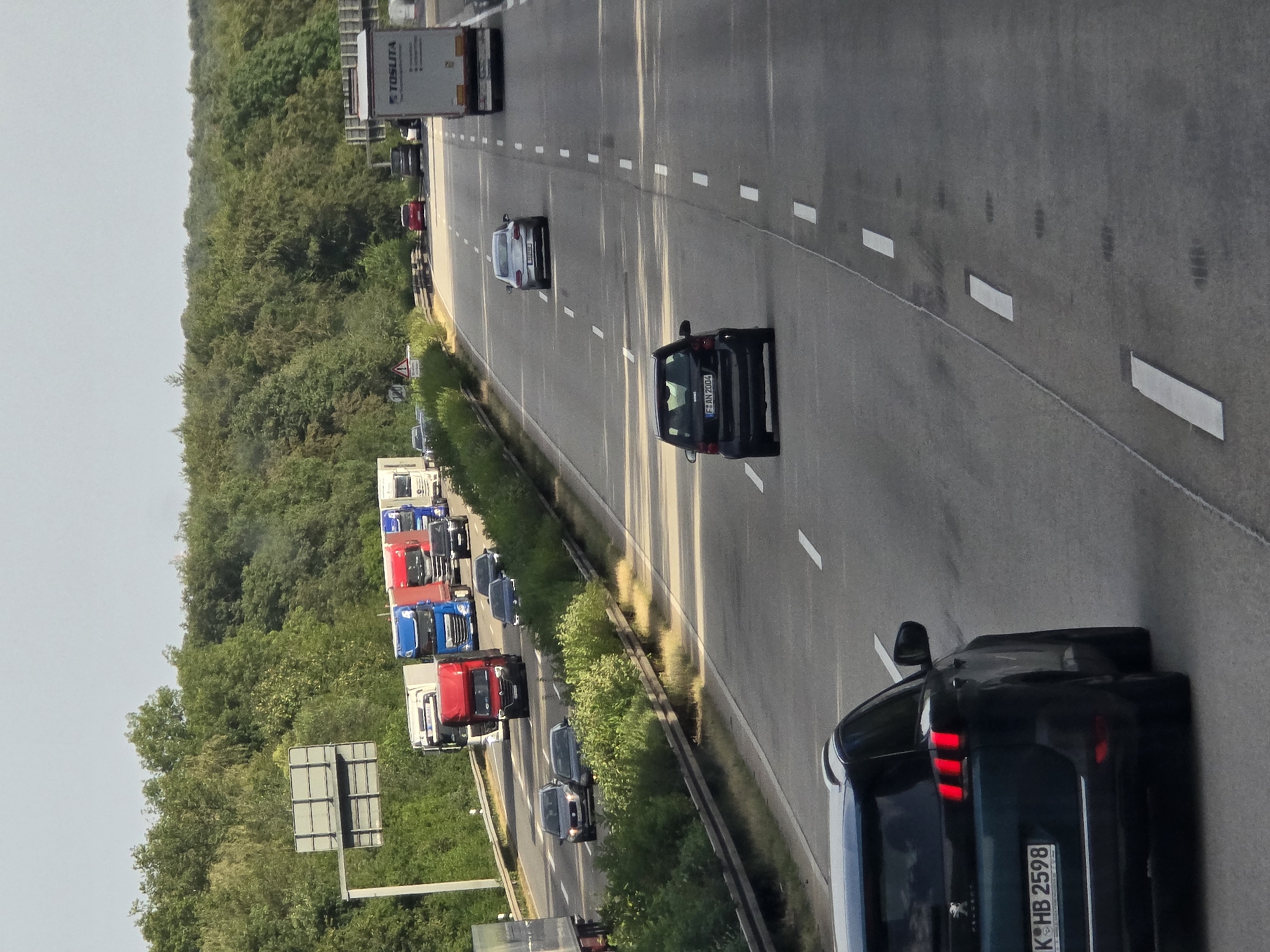 The image depicts a highway scene in Bremthal, Germany, with multiple vehicles, including trucks and cars, traveling in the same direction. The highway has multiple lanes, marked by white lines. Dense green trees line the sides of the highway, creating a natural backdrop. The sky is clear and overcast.
In the foreground, a dark-colored SUV is visible, its rear facing the camera. It has a license plate that reads "K-HB 2598". Further ahead on the highway, a black Smart car is seen with its license plate reading "F-AN 2004". A silver sedan is also visible, as well as several trucks, some of which are red and blue. A white truck with the logo "TOSLITA" is in the right lane. Traffic appears to be moderate. There are no visible pedestrians.