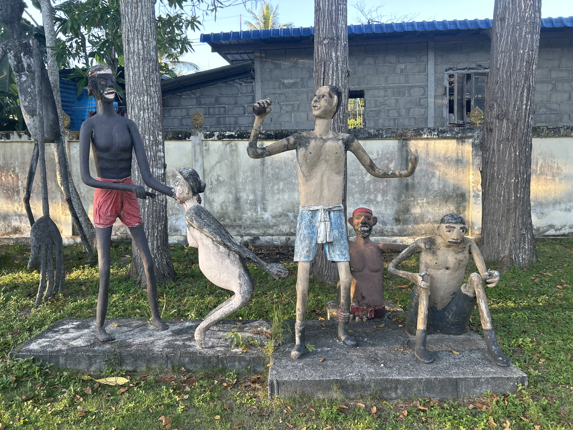 This image depicts an outdoor sculpture display in Ko Phangan, Thailand. The sculptures appear to be made of concrete and are situated on raised platforms in a grassy area. In the foreground, there are five figures. On the left, a dark-skinned male figure stands wearing red shorts and a headband, holding the arm of a lighter-skinned female figure who is bent over. To the right of the standing male figure, another dark-skinned male figure stands wearing blue shorts and a red headband. Next to him, a squatting dark-skinned male figure wears a red waistband. In the far right of the foreground, a seated male figure with dark skin wears dark shorts. The background features trees, a concrete wall, and a building with a blue roof. The lighting suggests it is daytime, possibly late afternoon given the shadows.