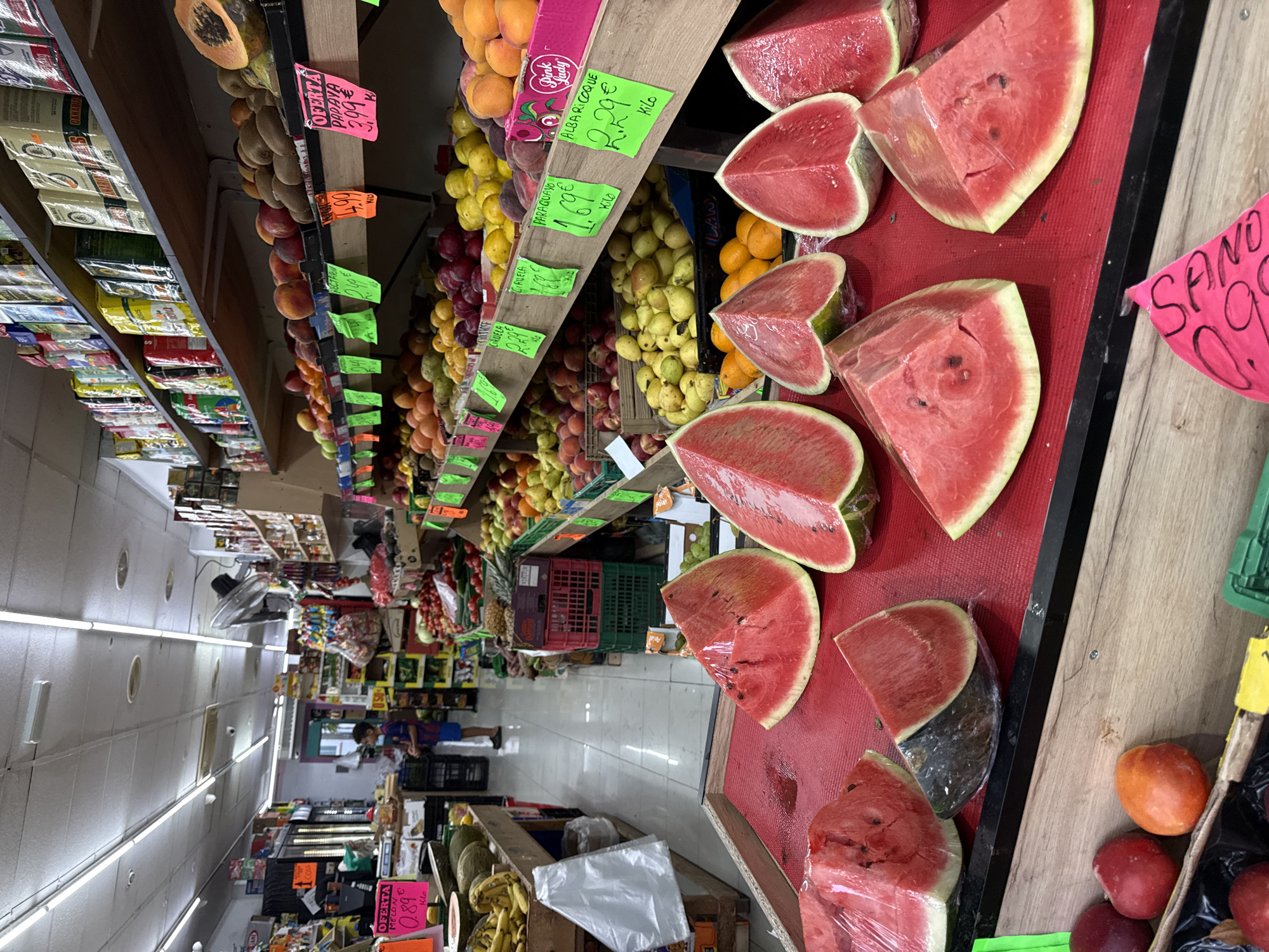 The image shows the interior of a fruit and vegetable market in Blanes, Spain. The market is well-stocked with a variety of fresh produce. In the foreground, several large slices of watermelon are displayed on a red mat, some covered in plastic wrap. Behind the watermelons, on a wooden shelf, are various other fruits including peaches, apricots, and yellow plums. Further back, more shelves are laden with kiwis, apples, and papayas. Cages filled with produce like tomatoes and cucumbers are visible on the right side of the image.
In the middle ground, a young boy in a blue t-shirt and blue shorts stands looking at the fruit. To his left, more produce is arranged on shelves, including bananas and melons. The market aisles are visible, with shelves stacked with packaged goods and boxes of fruit. Fluorescent lights illuminate the interior. The overall impression is that of a busy, well-organized local market.