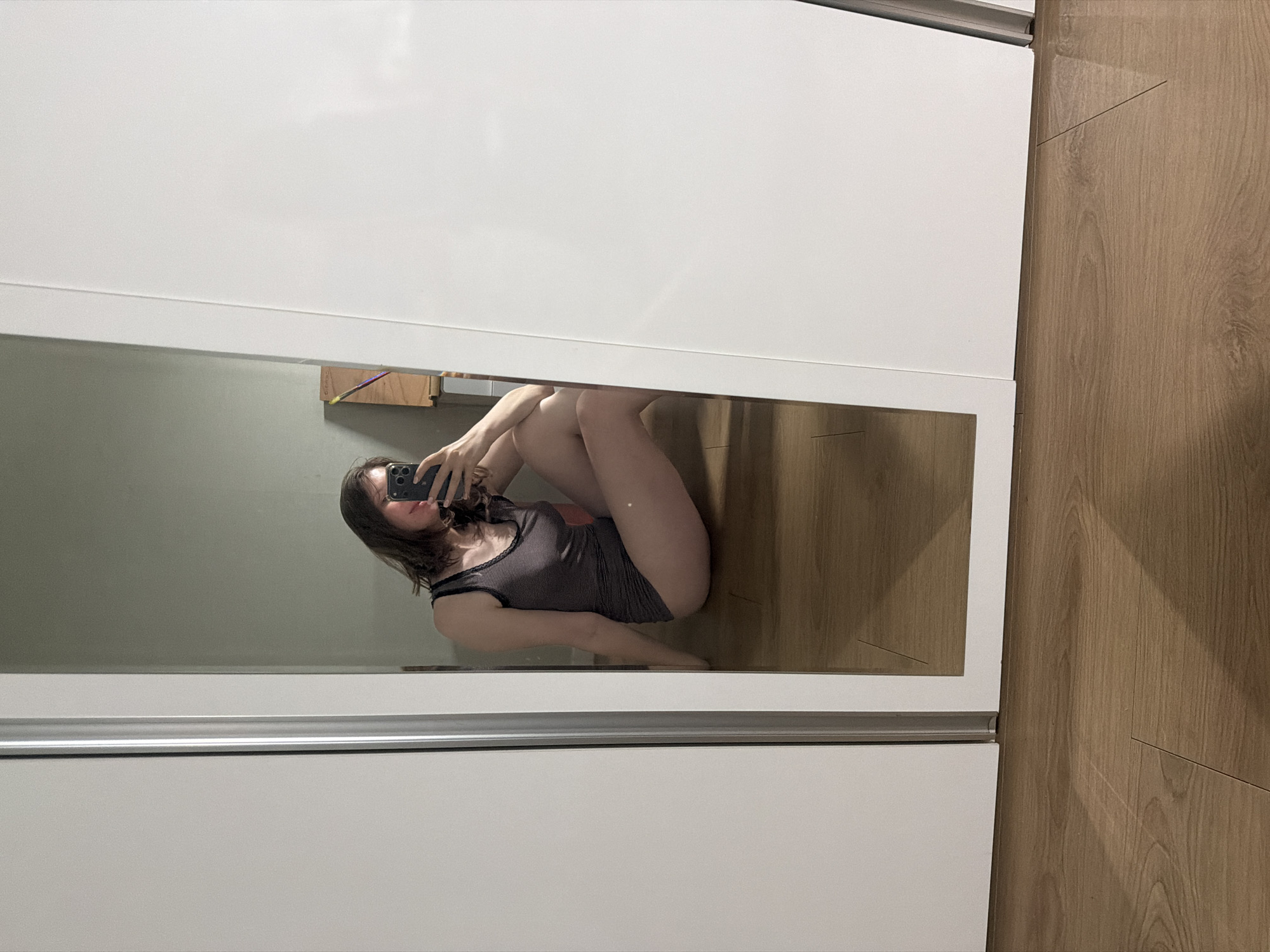 A young woman is captured in a mirror selfie, sitting on a wooden floor indoors. She is wearing a dark grey, sleeveless tank top with a lace trim. Her right arm is extended, holding a smartphone to take the picture, and her left arm is resting on her bent left leg. Her legs are crossed at the ankles, and she is leaning back on her hands. The background appears to be a room with a pale green wall, and a whiteboard or large white surface to her right. A closet with a silver track is visible to her left. The lighting suggests an indoor environment, possibly a home. The image does not contain any information that places it in Dongducheon-si, South Korea.