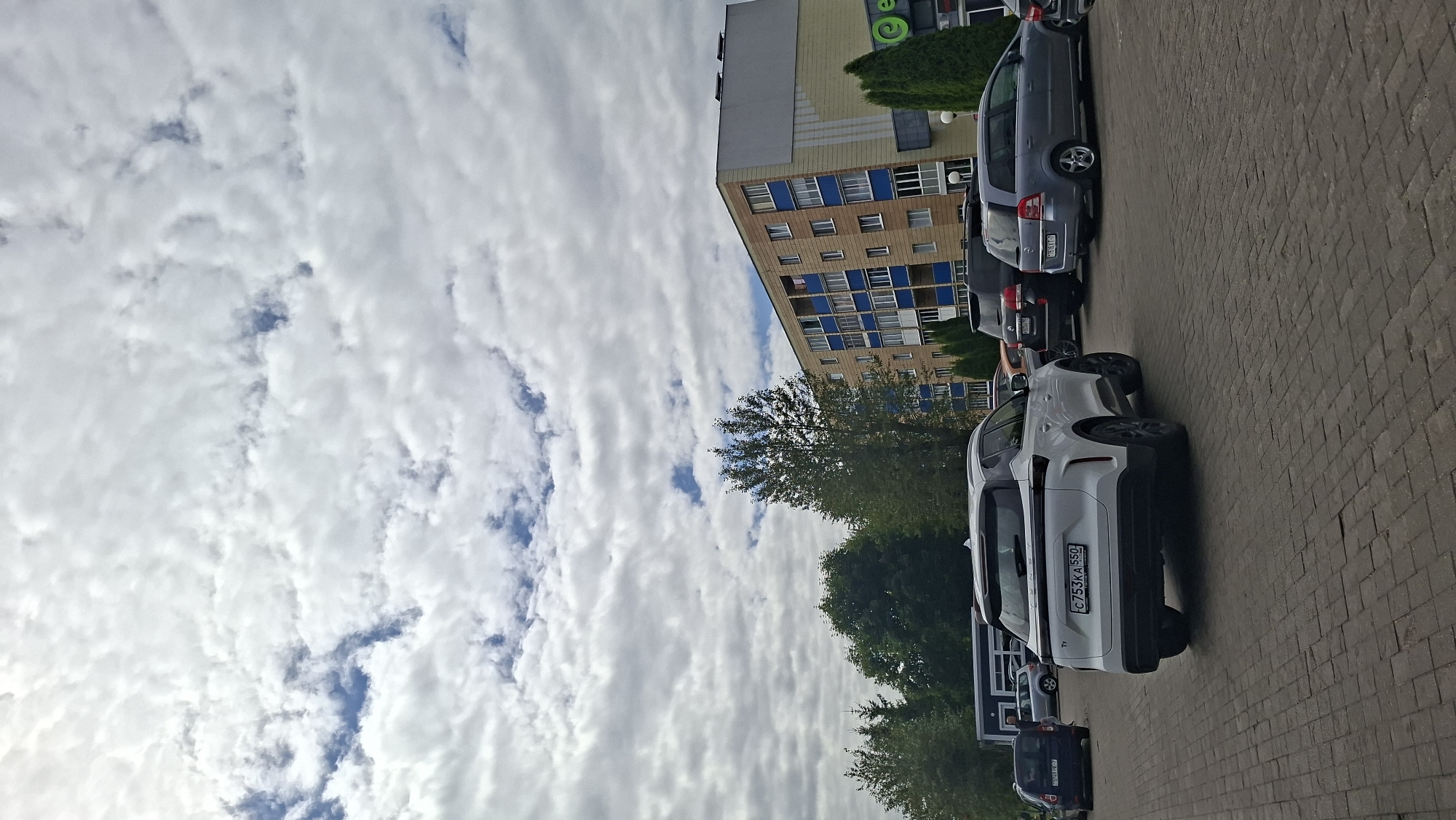 A white SUV is parked on a brick-paved lot in front of a multi-story building. The SUV is facing away from the camera, with its license plate visible, showing "C753KA 550". Other vehicles are parked nearby, including a dark-colored SUV and a gray station wagon. The building appears to be an apartment complex or commercial building with balconies and blue accents. Trees and a cloudy sky are visible in the background. The scene is likely in Myadzyel, Belarus, given the Cyrillic license plate and architecture.