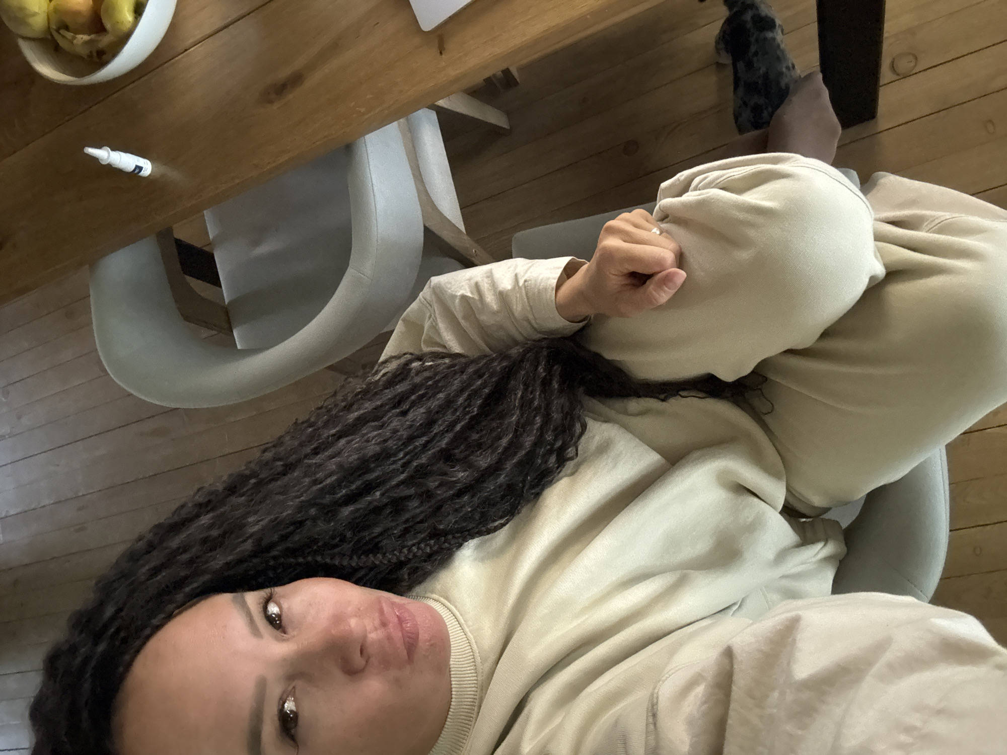 An overhead shot captures a woman relaxing indoors, likely in Chaikovsky, Russia. She is seen from above, resting in a light grey upholstered chair at a wooden table, wearing a comfortable light beige sweatshirt and matching sweatpants. Her head is tilted, and she gazes directly up at the camera with a neutral, slightly pouty expression. Her long, dark, braided hair flows over her shoulder and chest. A silver ring is visible on her right ring finger.
On the wooden table, to her left, sits a white bowl containing yellow or orange fruits, possibly lemons or oranges. Nearby, a small white bottle, resembling eye drops, with an unreadable blue label is present. The flooring is made of wooden planks. In the background, partially visible under the table near her left foot, is a patterned grey and black dog. The bright, even lighting suggests it is daytime. The overall scene conveys a sense of casual comfort and domestic relaxation.