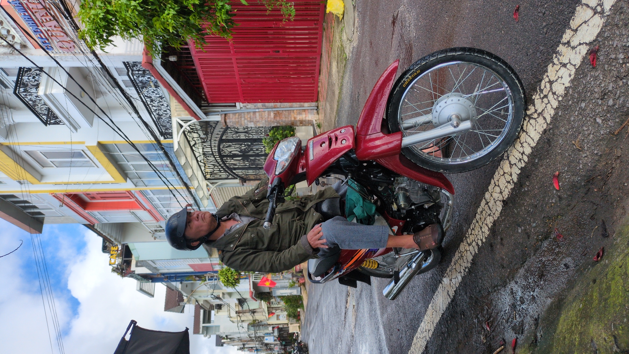 A middle-aged East Asian man wearing a dark helmet and a green jacket is seated on a red motorbike. He is wearing gray pants and brown sandals. The motorbike is parked on a wet asphalt street, indicated by puddles and a white painted line on the road. The street is lined with multi-story buildings with colorful facades, including yellow, red, and white. Some buildings have decorative iron balconies and red fences. A lush green tree is visible on the right side of the image. The sky is cloudy with patches of blue. This scene appears to be in Da Lat, Vietnam, a city known for its French colonial architecture and its cool climate, which often results in wet streets. The presence of various architectural styles and the general street scene are consistent with urban areas in Da Lat.