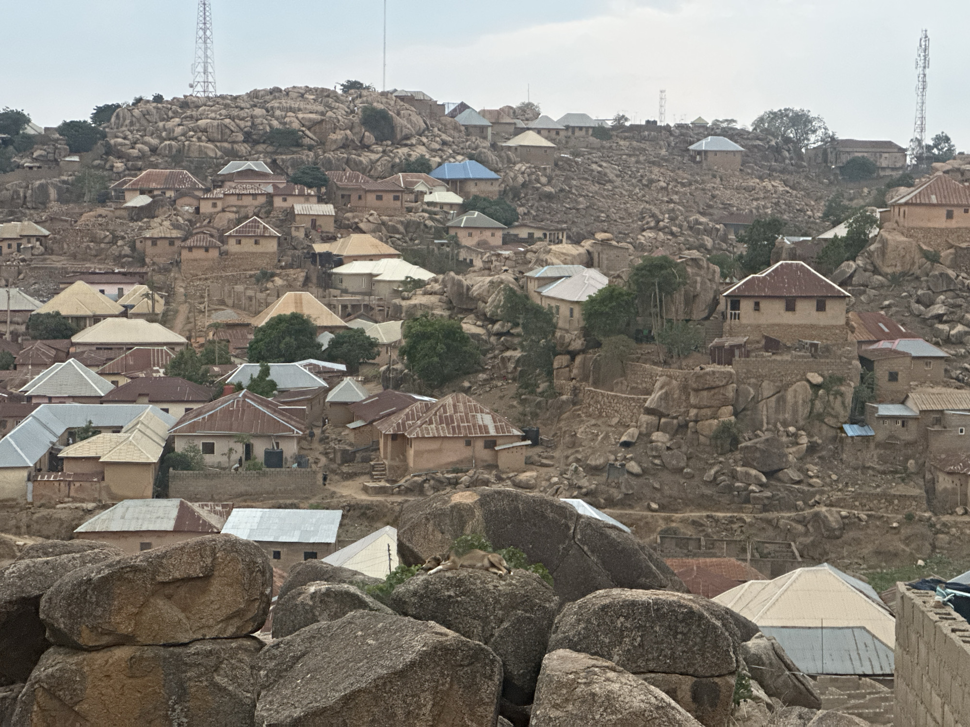 The image depicts a densely populated, rocky landscape in Jos, Nigeria. Numerous houses are nestled amongst and built upon large boulders, creating a tiered settlement. The structures have a variety of roof colors, including blue, white, and red-brown. Several cell towers are visible in the background. In the foreground, large boulders dominate the view, with a dog resting on one of them. No people are clearly visible, but their presence is implied by the density of housing and the paths traversing the rocky terrain. The overall environment appears arid and dusty.