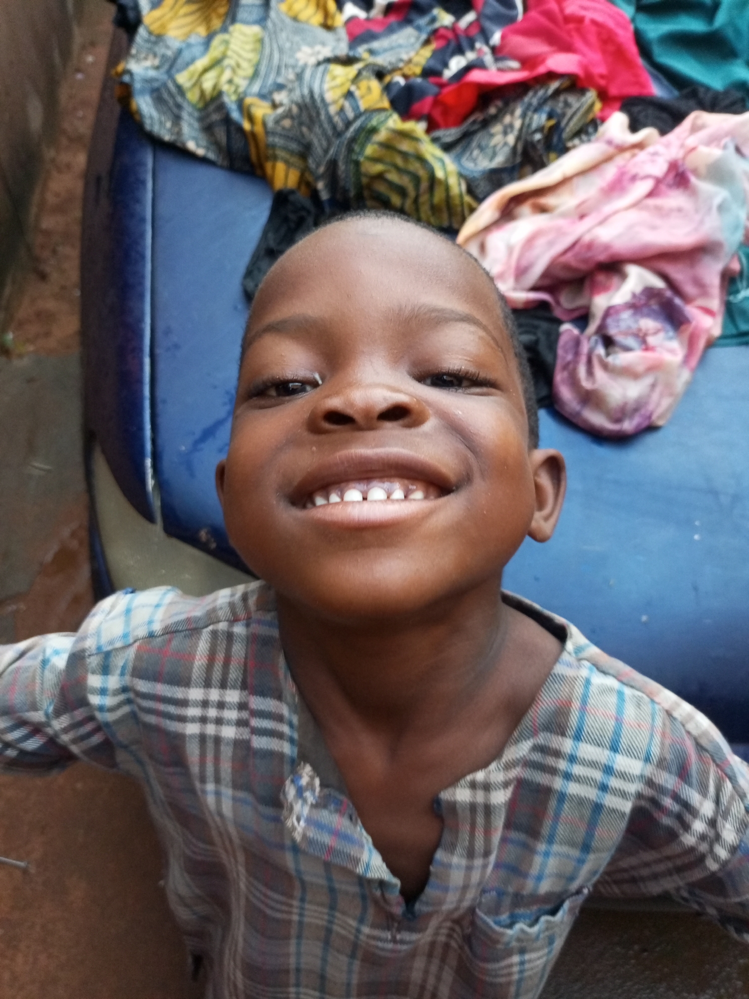 A young Black child, likely a boy, is shown in a close-up portrait with a bright smile. The child has dark skin, dark eyes, and short, dark hair. They are wearing a plaid shirt with colors of grey, blue, and red. The child's mouth is open, revealing their teeth. In the background, there are colorful fabrics piled on what appears to be the blue surface of a vehicle. The overall impression is one of joy and innocence. The location is unknown.