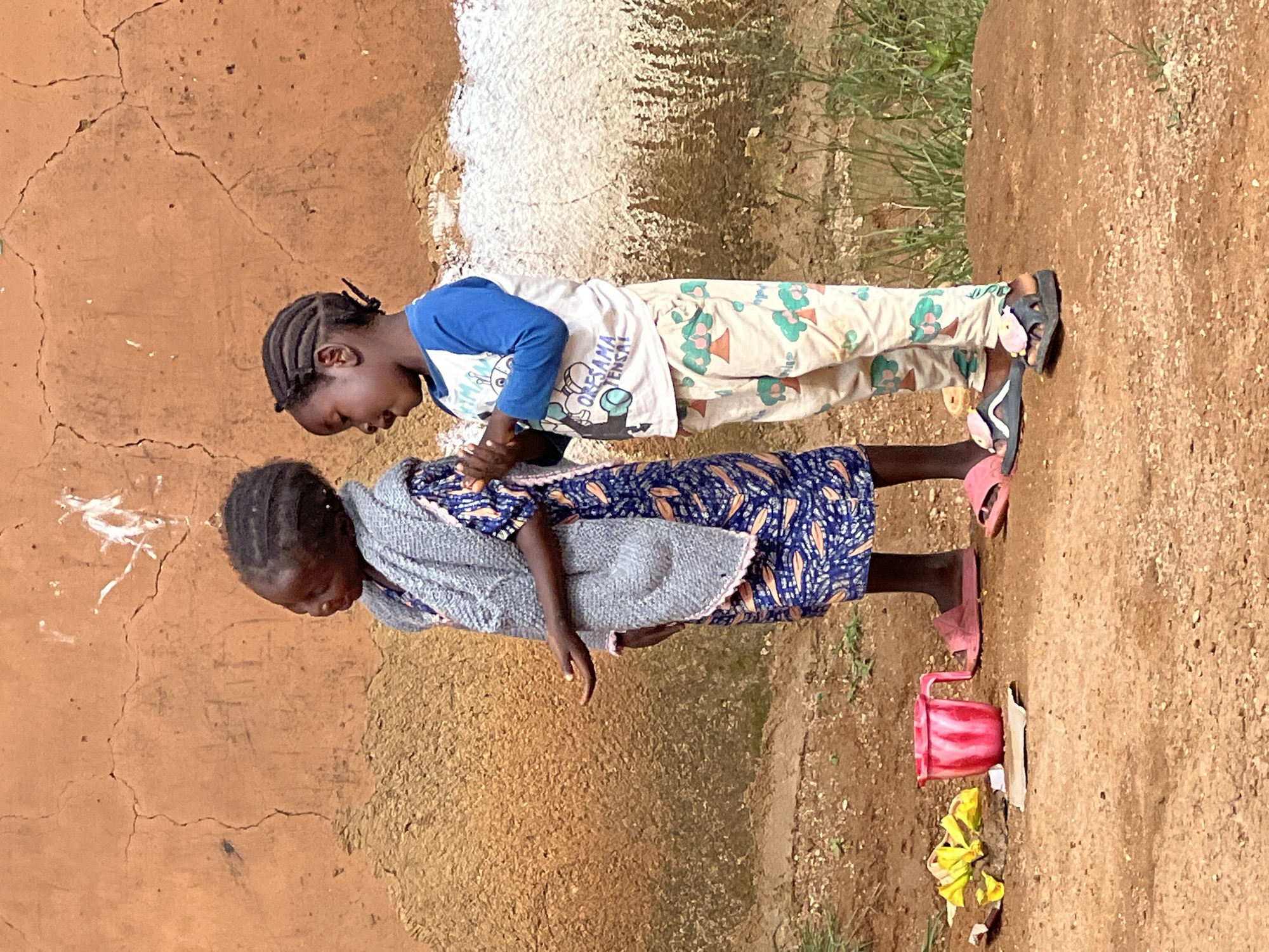 Two young girls stand next to a rough, orange-colored wall in Jos, Nigeria. The girl on the left wears a blue dress with an orange pattern and a grey shawl. She has her left hand extended, palm up, as if asking for something. The girl on the right wears a blue long-sleeved shirt and patterned pants with images of trees. Her hair is braided and pulled back with a dark elastic. She appears to be speaking or singing. In front of the girls on the ground, there is a pink plastic cup, some yellow flowers, and a piece of cardboard. The ground is made of dirt and small stones, with some patchy green grass growing in the background. The wall behind them is textured and cracked, with some white and dark markings.