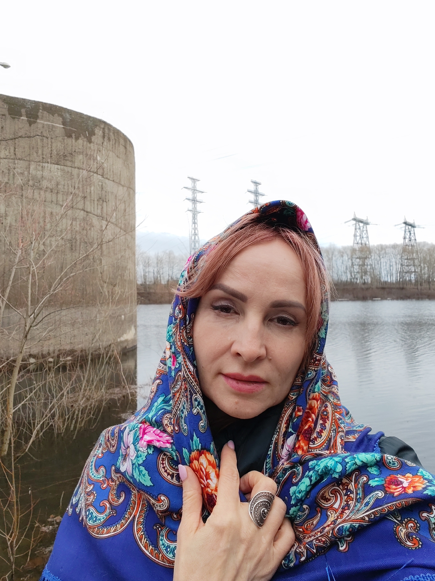 A woman is posing for a selfie in front of a concrete structure by a body of water, likely the Kama River. In the background, electrical pylons are visible. The woman is wearing a blue headscarf with a floral pattern, a black jacket, and a large silver ring on her hand. The location appears to be near a reservoir or power plant on the edge of Chaikovsky, Russia. The overall impression is one of a damp or cold day, given the gray skies and leafless trees.