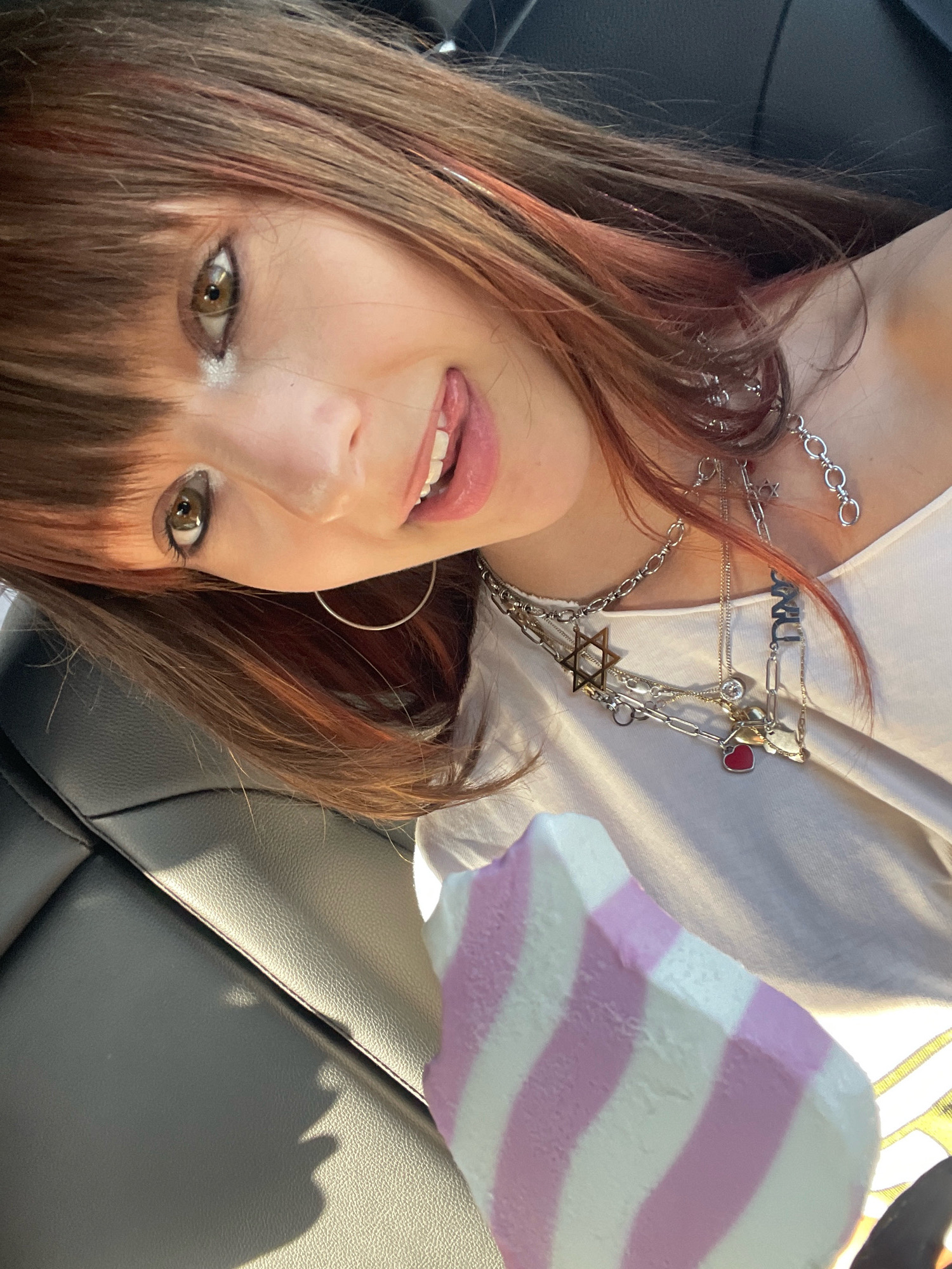 The image shows a close-up, slightly tilted selfie of a young woman with brown hair with pink streaks, who is sticking her tongue out at the camera. She is wearing a white top and multiple silver necklaces, including one with a Star of David pendant and a red heart charm. She has on dark eye makeup. In the foreground, a portion of a striped pink and white object, likely an ice cream bar, is visible. The background suggests an indoor setting, possibly a car, with a textured black surface visible on the left, likely leather seating. The lighting indicates it is daytime, with a strong light source casting shadows. The image does not contain any discernible features that would indicate a specific location within the city of Raanana, Israel.