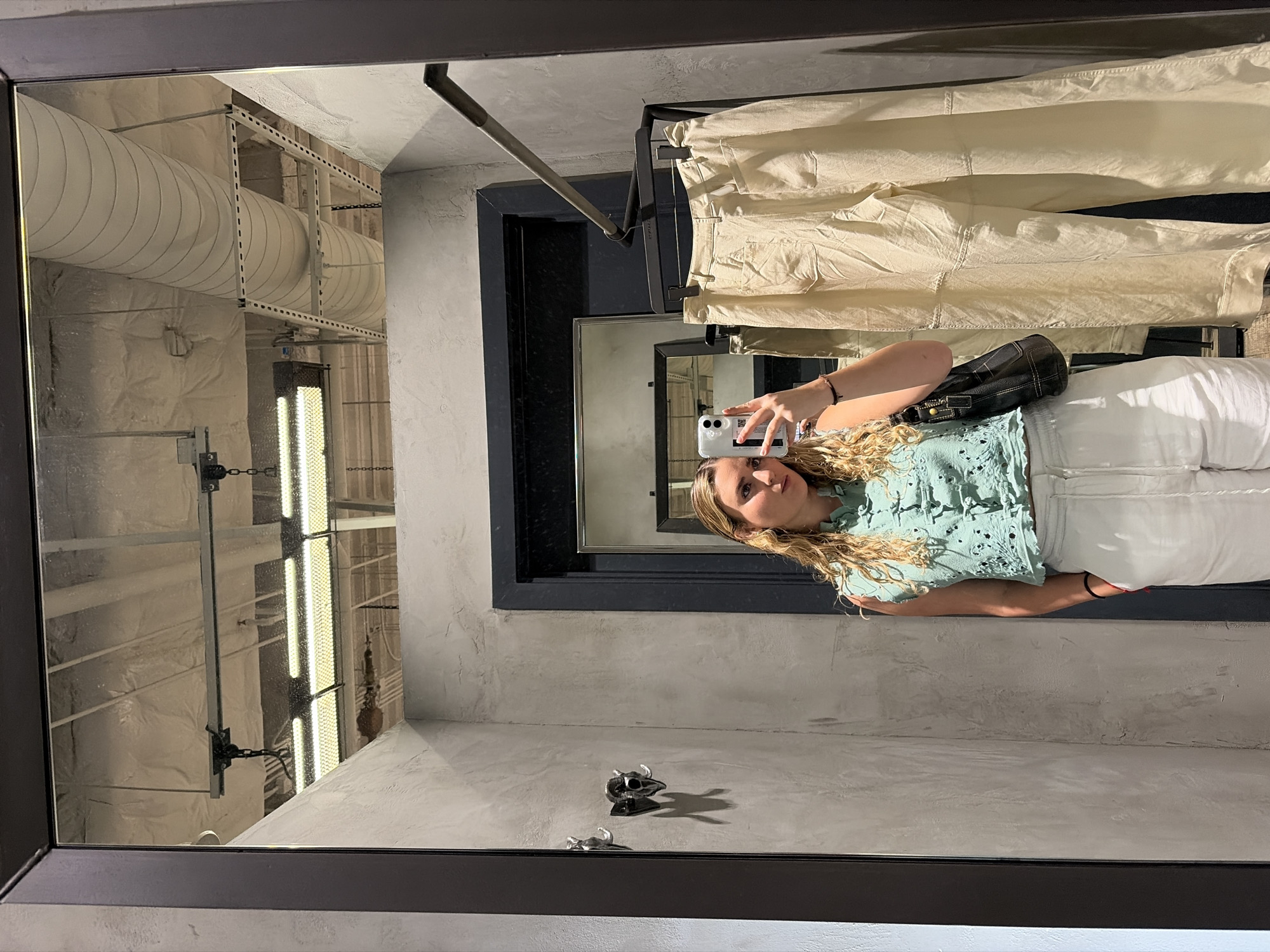 A person is taking a photo of themselves in a mirror, which is likely located in a store's changing room. The person is wearing a light green, textured top and white pants. They are holding a white smartphone in their right hand. A black purse is visible on their shoulder.
The changing room has a metal rack above with light-colored clothing hanging from it. Behind the person in the mirror, another mirror is visible, reflecting a lighting fixture and a ceiling that seems to have a commercial or industrial design with large air ducts. The walls of the changing room appear to have a gray textured finish. On the base of the mirror, small animal-like figurines or decorations are seen. The mirror has a dark-colored frame.
Given the attire of the person and the general appearance of the changing room, the location is likely a clothing store. The presence of industrial-style ceilings and air ducts suggests the possibility of a renovated or repurposed building, which can be found in various parts of Miami. Further clues, such as specific store details or landmarks reflected in the mirror, would be necessary to confirm the exact location within Miami.