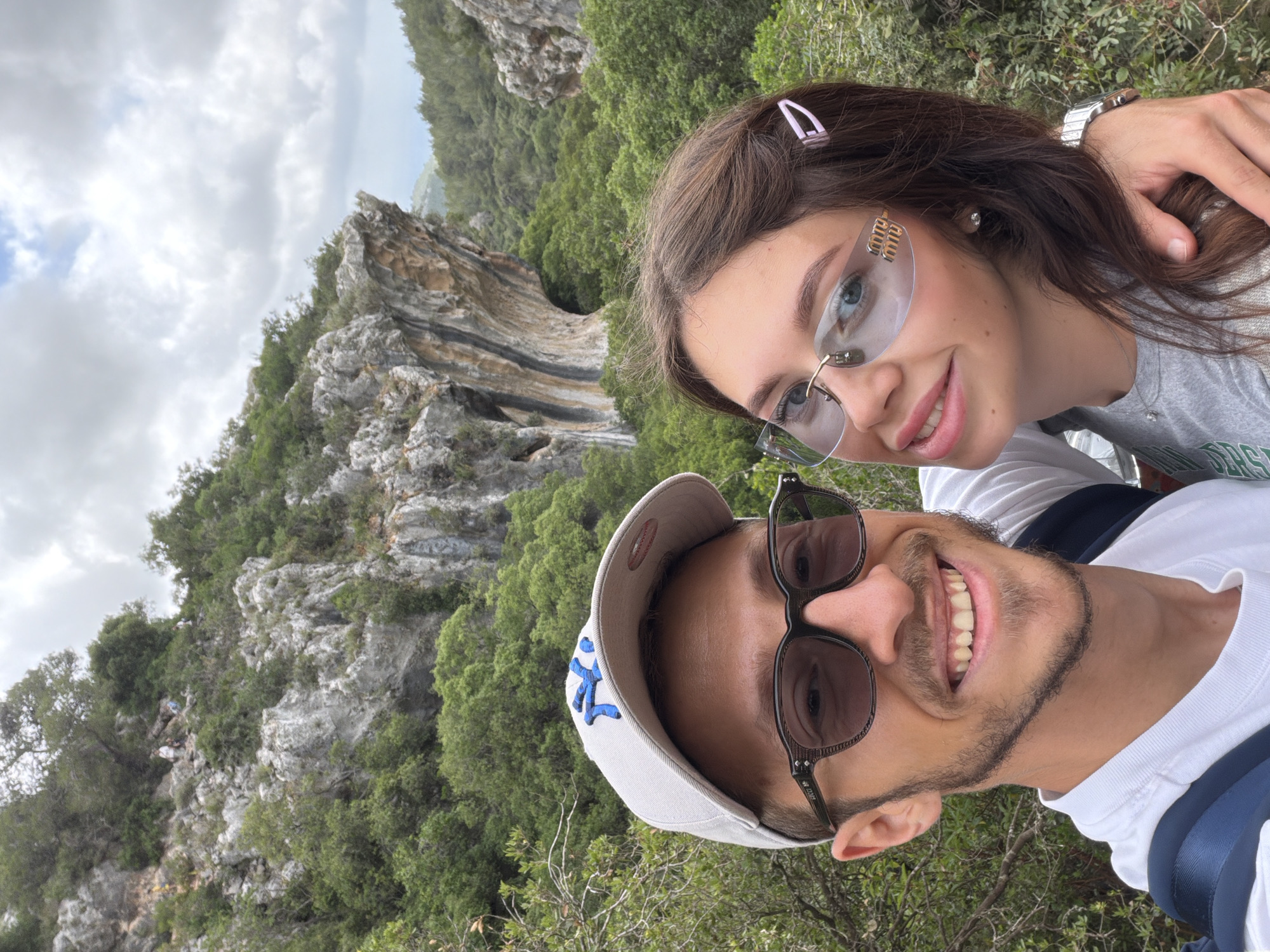 A selfie taken outdoors in Tirat Karmel, Israel, captures a smiling man and woman. The man wears sunglasses and a baseball cap. The woman wears sunglasses and a hair clip, with a wrist watch visible on her left wrist. They stand in front of a rocky landscape with trees and cloudy sky in the background.
