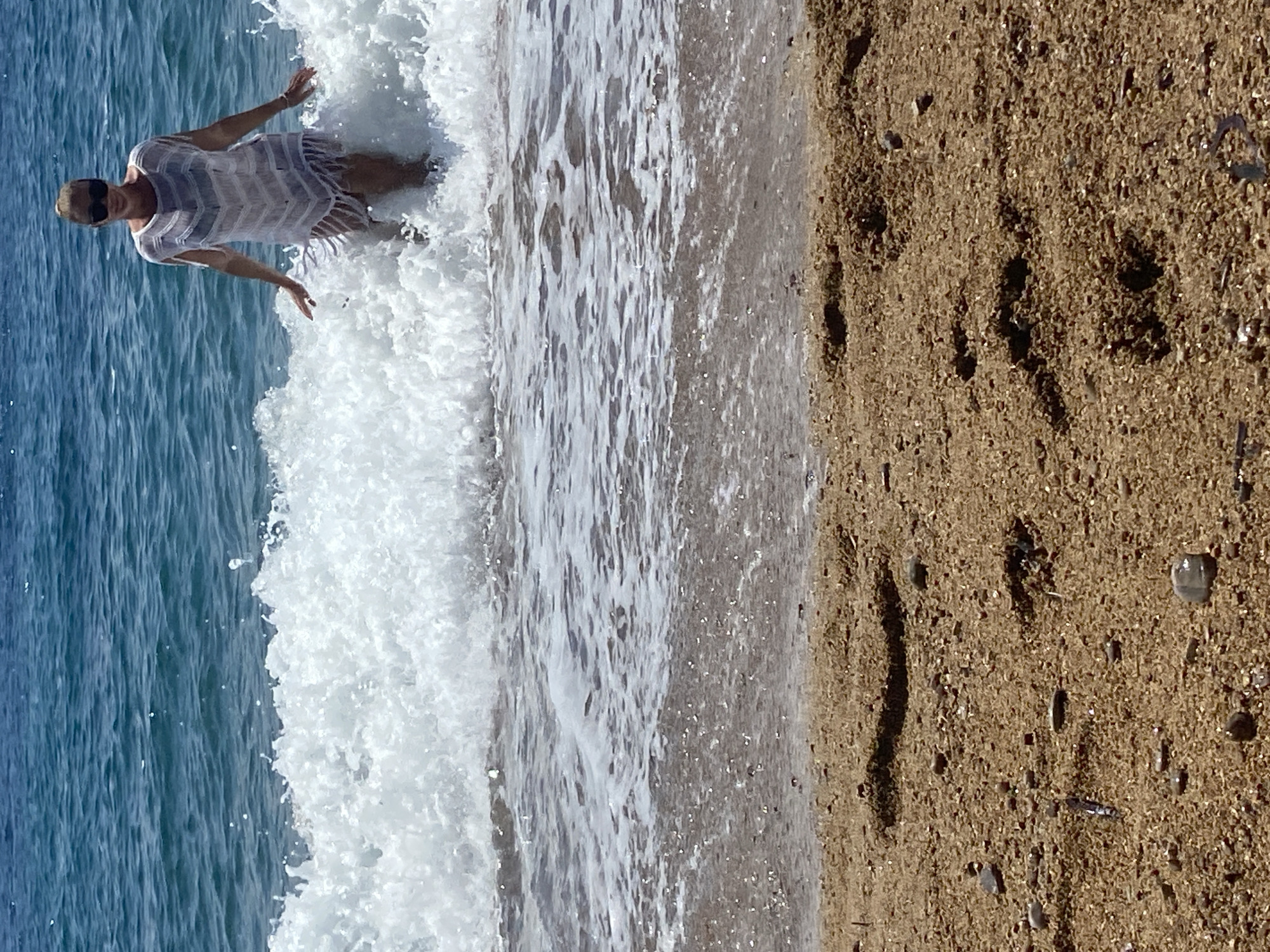 An outdoor, eye-level shot captures a woman wading in the shallow waters of a beach, set against the backdrop of the sea. The image is described as taking place in Sveti Stefan, Montenegro.
The main subject is a woman, seen from the waist up, standing amidst a white, breaking wave. She wears dark sunglasses and a light-colored, possibly white or off-white, sheer or open-knit cover-up or dress, which is wet from the splashing water. Her light-colored hair is pulled back. Her arms are slightly outstretched and bent at the elbows, with open hands, suggesting a moment of enjoyment, balance, or embrace of the incoming wave.
To the left of the woman, the deeper blue-green ocean stretches out, showing subtle ripples on its surface. To her right, the light brown sand of the beach is visible, textured with small pebbles and grains. The foamy white water of the wave extends from the open sea to the sandy shore, creating a clear line between the two.
The scene is brightly lit, indicating a sunny daytime with clear weather. The overall impression is one of leisure and pleasure by the sea. There is no visible text in the image.