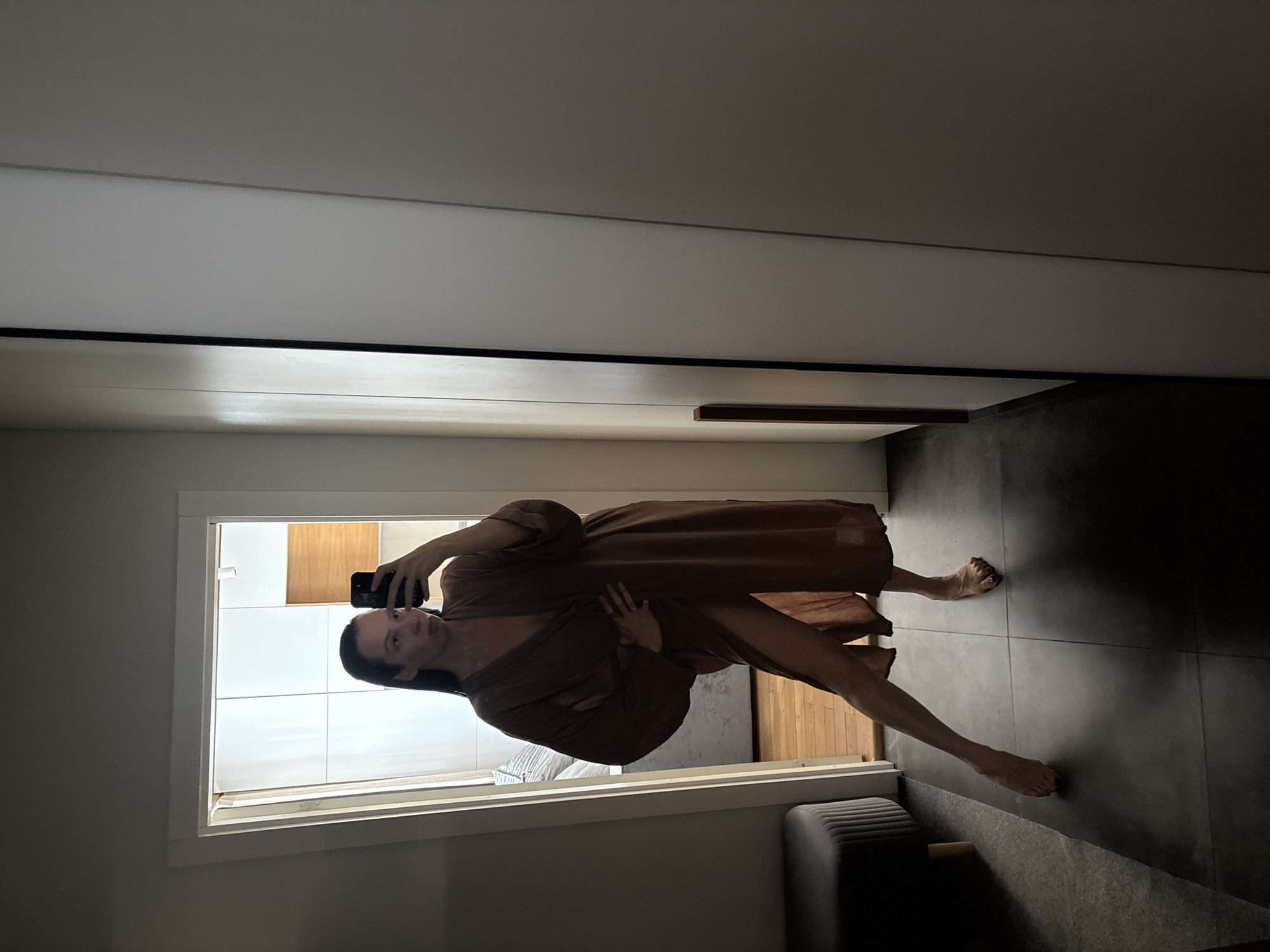 A woman in a brown robe takes a selfie in a modern bathroom. The bathroom has dark grey floor tiles and a white door. There is a grey ottoman in the corner. The bathroom in the reflection has white wall tiles and wooden cabinets. This image was taken indoors. The location is the city of Chaikovsky, Russia.