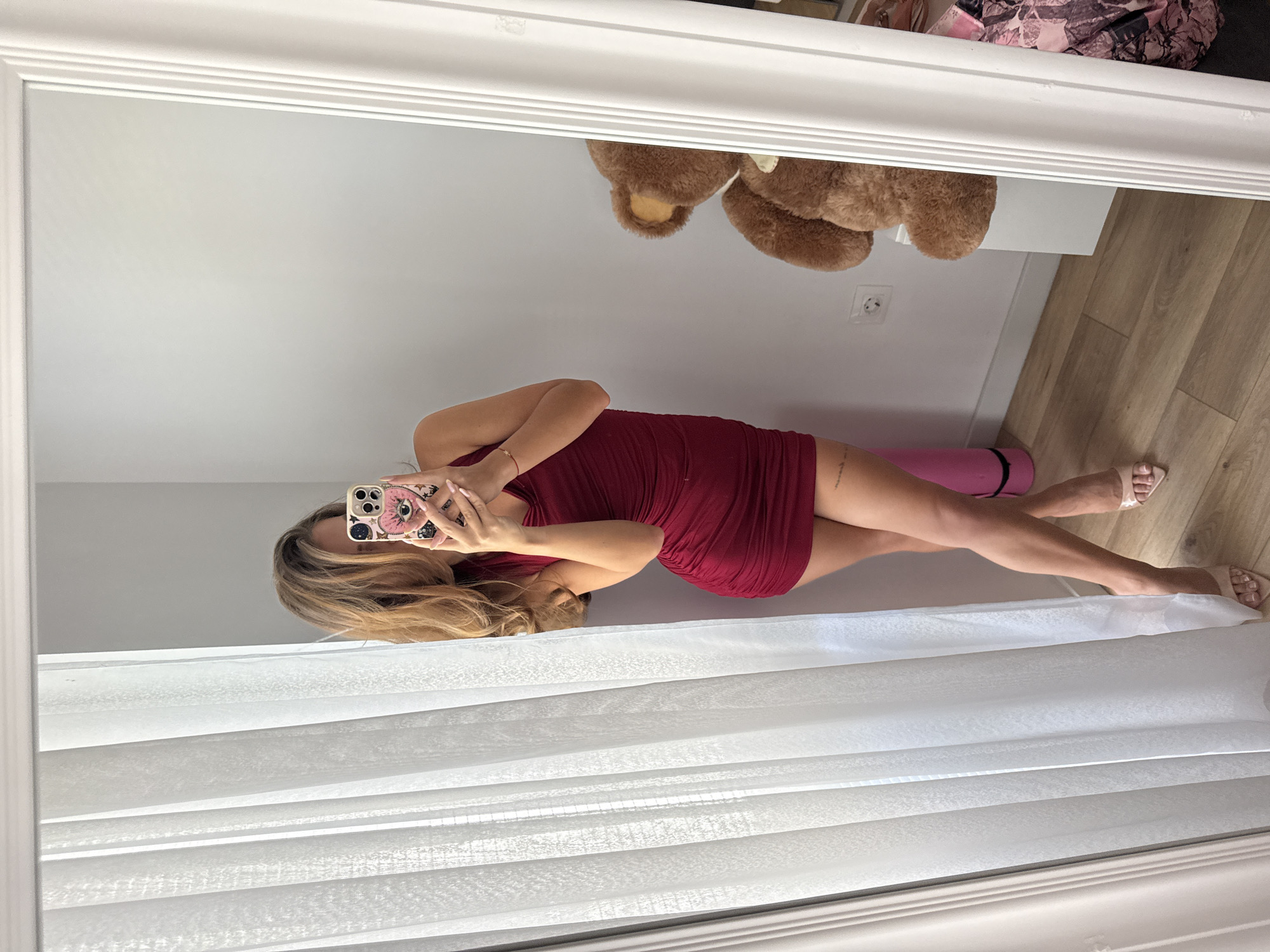 A young woman with blonde hair is taking a selfie in a full-length mirror, wearing a short, form-fitting red ruched dress and nude heeled sandals. Her phone case has a pink heart with an eye design. She is standing next to a white sheer curtain. In the reflection of the mirror, a large brown teddy bear is visible on a white shelf, along with a pink yoga mat leaning against the wall. The floor is made of light-colored wood. The room appears to be a domestic setting, possibly a bedroom or dressing area, located in Plovdiv, Bulgaria.