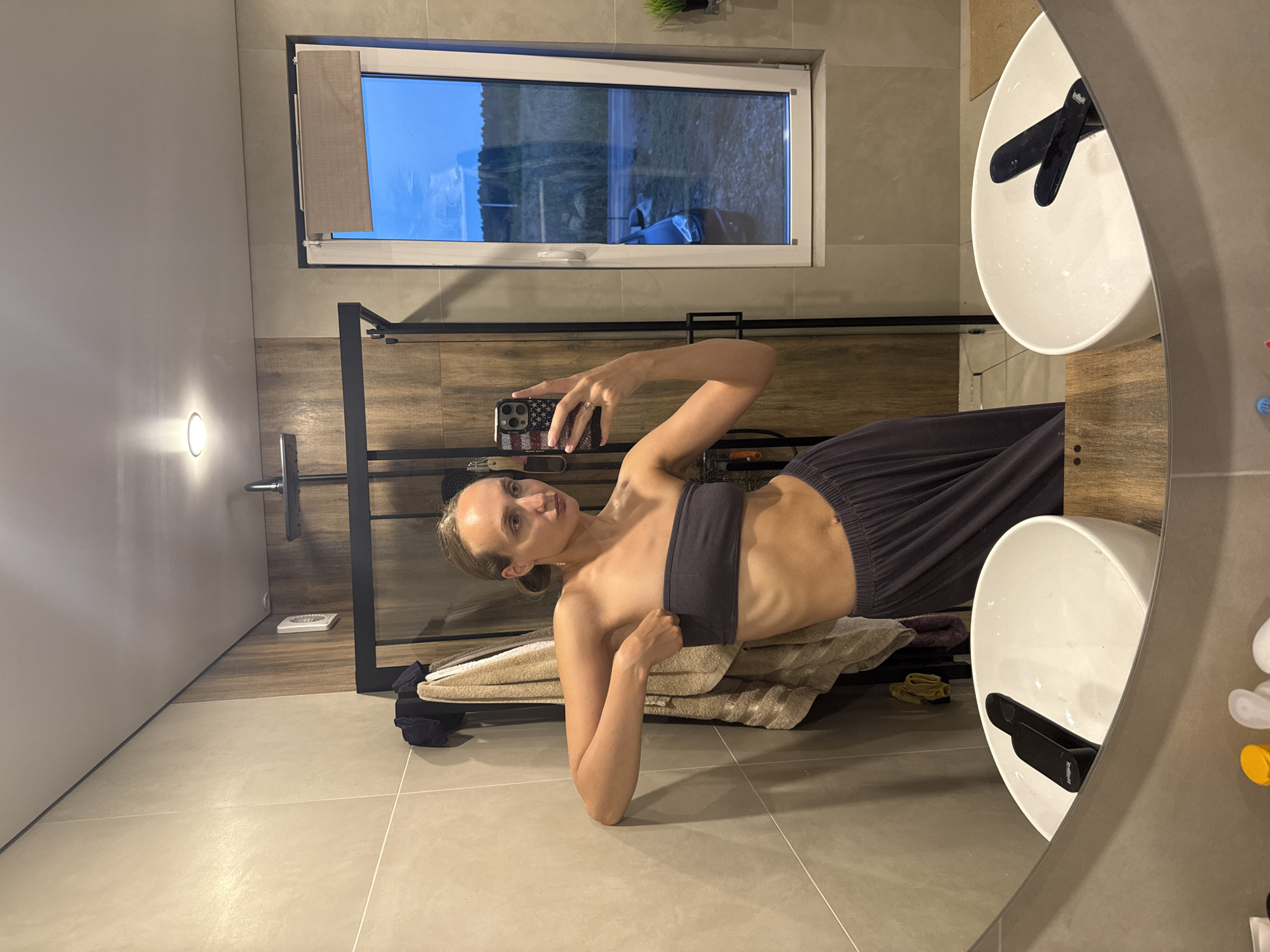 A woman stands in a bathroom, posing for a selfie in a mirror. She is wearing a dark grey strapless top and matching wide-leg pants. Her left arm is leaning against the tiled wall. In the background, a glass-enclosed shower with wooden accents is visible. To the right of the shower is a window offering a view of an outdoor scene with a car. Two white sinks with black faucets are in the foreground, reflected in the mirror. The overall impression is that of a modern, well-lit bathroom. The location is identified as Podshivalovo, Russia.