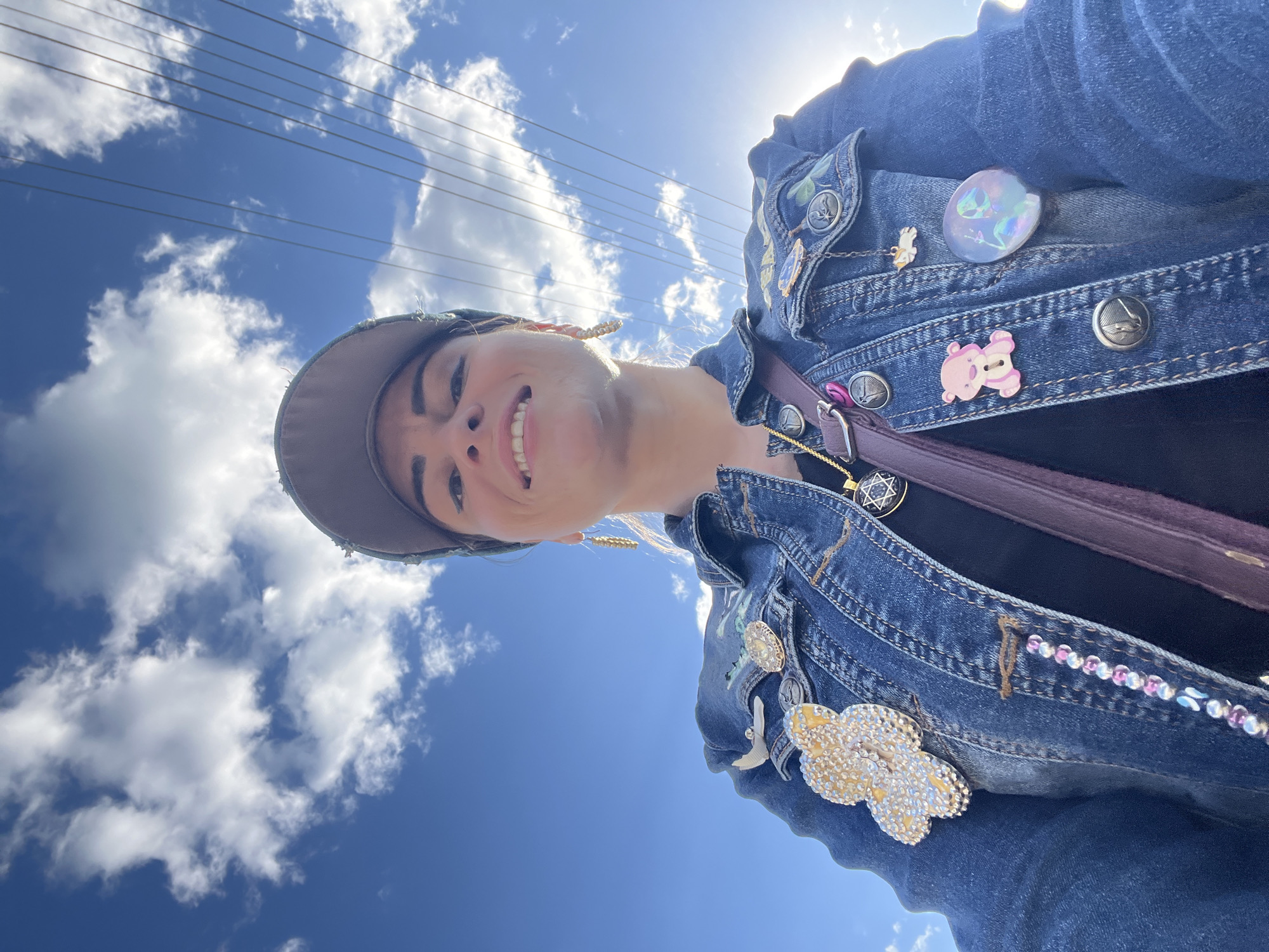 The image is a close-up, low-angle selfie taken outdoors on a sunny day. The primary subject is a person wearing a denim jacket adorned with various pins and embellishments. The pins include a pink teddy bear, a clear oval iridescent pin with iridescent figures inside, a small white bird, and a large sparkly cloud-shaped pin. There is also a beaded embellishment along the edge of one of the jacket's pockets. The person is wearing a baseball cap, and their face is visible, smiling at the camera. Gold earrings and a necklace with a pendant featuring a Star of David are also visible. The background is a bright blue sky with fluffy white clouds and power lines running horizontally across the frame. The sun is visible and casting a lens flare. The overall impression is of a casual, cheerful moment outdoors, possibly in Krasnoe-na-Volge, Russia, given the location context. The time of day appears to be during daylight hours, likely mid-morning or afternoon, with clear, pleasant weather.