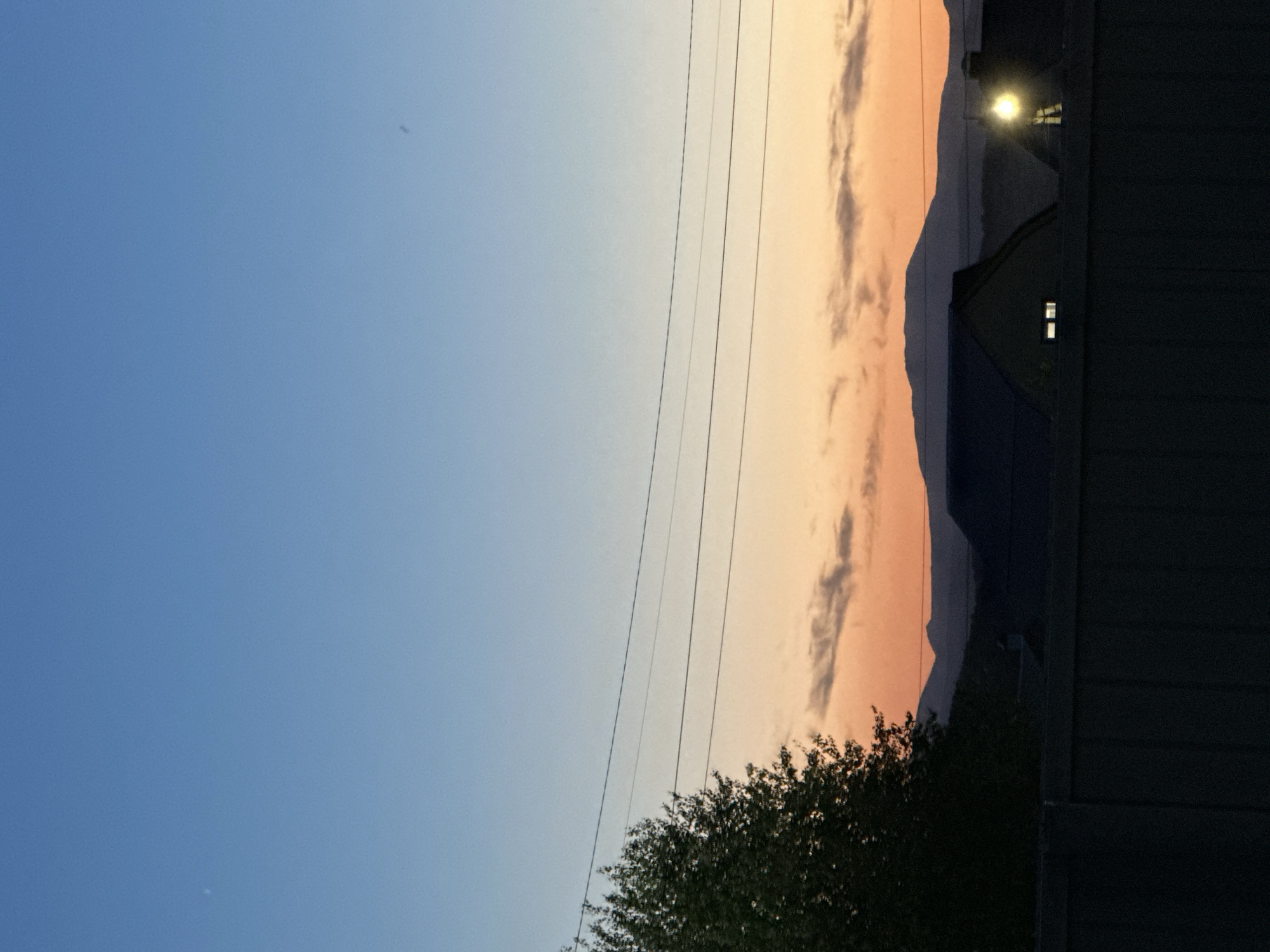 The image shows a sky with a gradient from blue to orange. Power lines are visible in the middle. In the background, there are dark silhouettes of mountains and a building with a lit window and a bright light above it. In the foreground, there's a dark bush and the side of a dark building.
