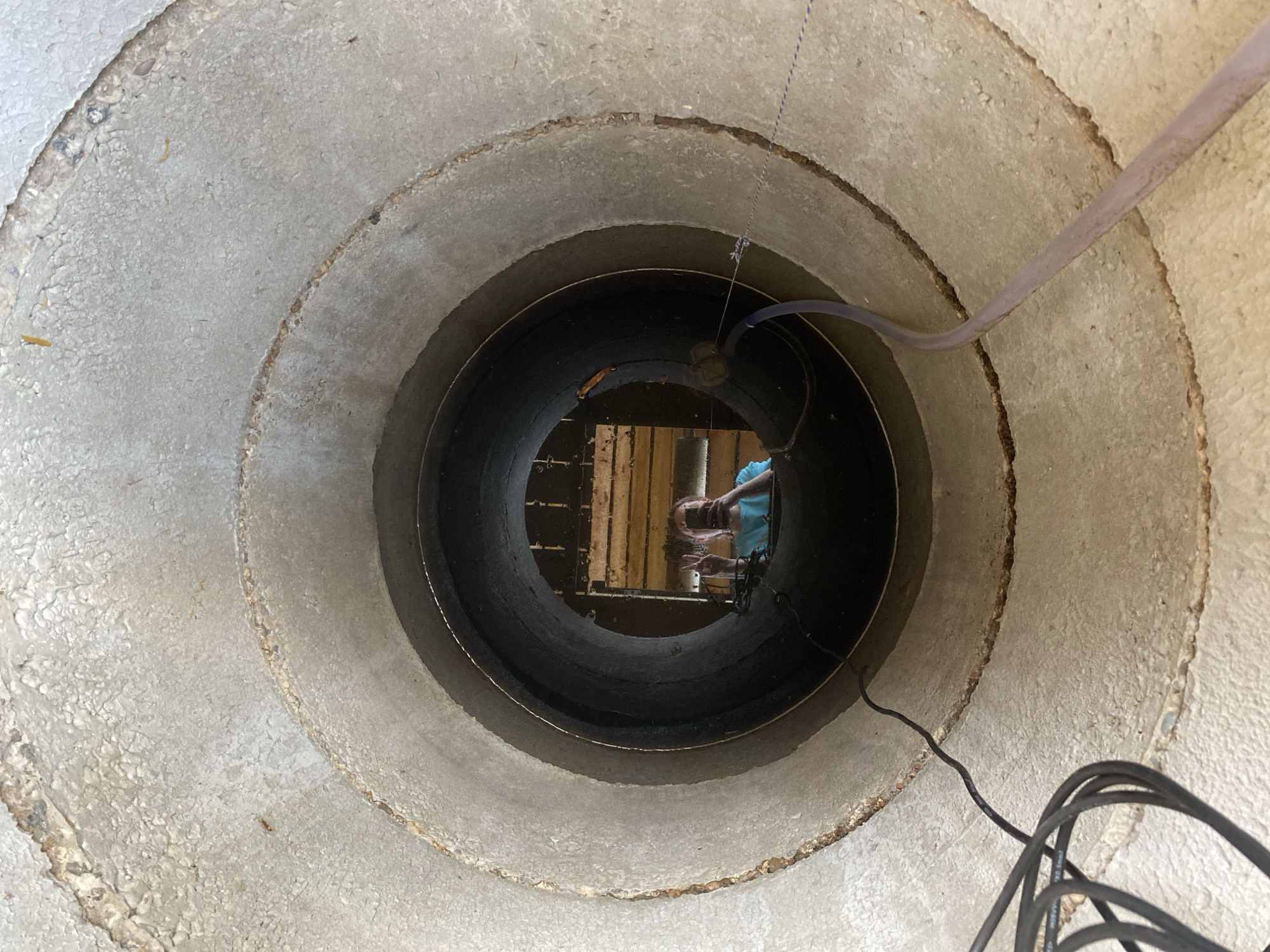 A circular, concrete well is viewed from above, revealing a dark, stagnant pool of water at the bottom. Reflections on the water's surface suggest a person leaning over the well, possibly looking down. Wooden beams, possibly a makeshift structure or debris, are partially submerged in the water. A grey hose and a black electrical cable extend down into the well. The concrete walls of the well are layered, showing signs of wear and age. There is no information about the location in the city of Chaikovsky, Russia from the provided image.