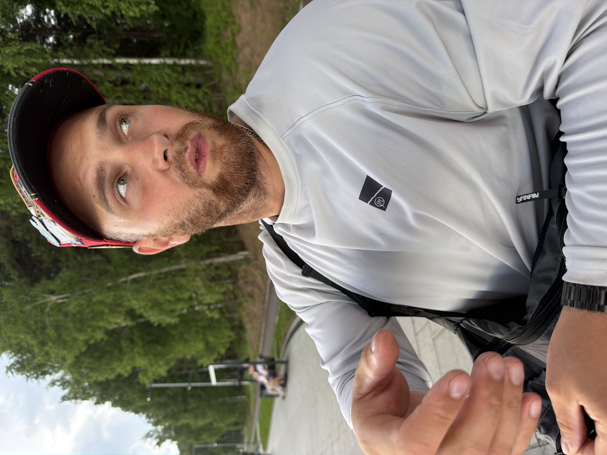 A close-up medium shot shows a Caucasian male in a light grey long-sleeved shirt and a red and black baseball cap, looking off to the right. He has light skin, green eyes, a short red beard, and is wearing a silver chain. His left arm is bent with his hand raised, and his right hand is visible near his waist. A black crossbody bag is slung over his right shoulder. In the background, blurred trees line a paved walkway with lampposts. Further in the background, two people are seated on a bench. The setting appears to be an outdoor park or recreational area in Chaikovsky, Russia, potentially along the Kama River embankment, given the typical landscape and urban planning of such locations in the city. The sky is overcast.