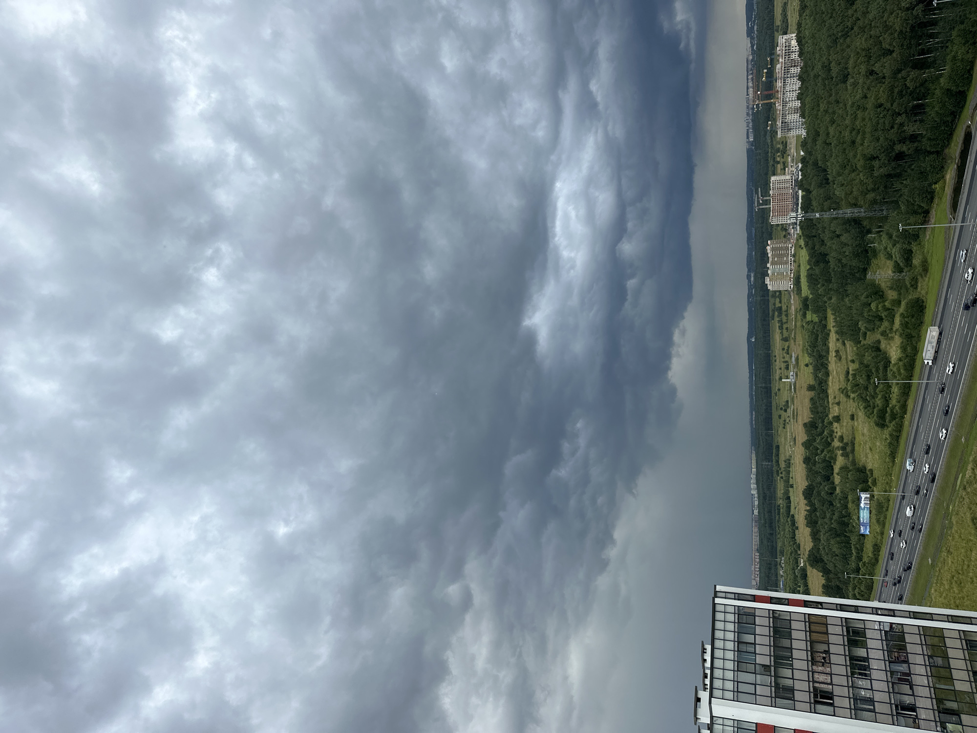 This image shows a cloudy sky over a landscape with a highway and buildings. In the foreground on the left is a tall, modern apartment building with many windows. To the right of the building is a multi-lane highway with cars and trucks traveling on it. Beyond the highway is a vast expanse of green fields and trees. In the distance, there are several large buildings under construction and some residential buildings. The sky is filled with heavy, dark clouds, suggesting an approaching storm. The location is identified as Novosergievka, Russia. There are no people visible in the image.