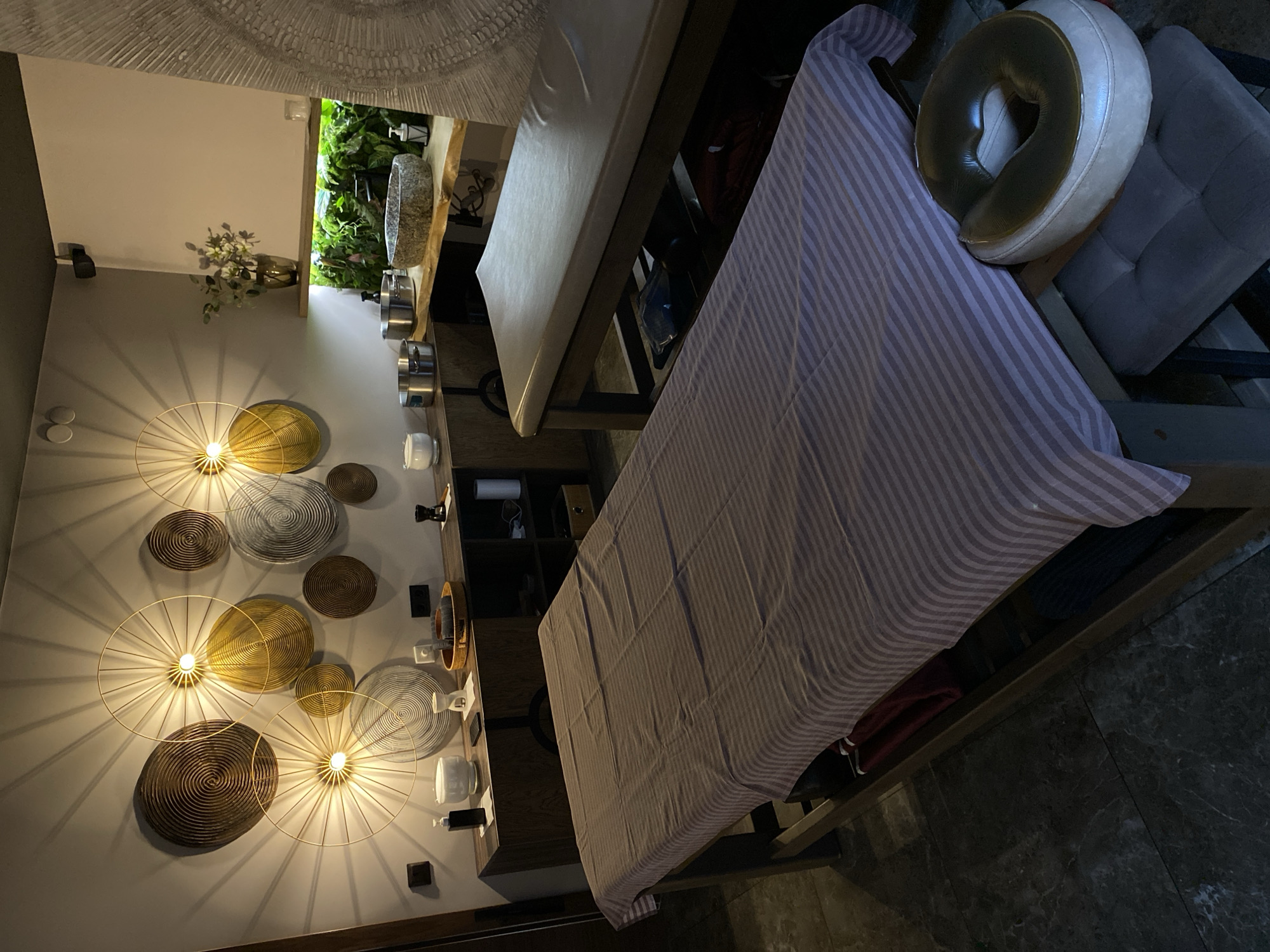 The image depicts a room, potentially a spa or wellness center, with a focus on a massage table covered in a striped sheet. A headrest is visible at one end of the table. The walls feature decorative lighting elements and wall-mounted shelves or cabinets. The overall color palette is warm and inviting. It is not possible to determine if this image is in the city of Votkinsk, Russia or any other specific location.