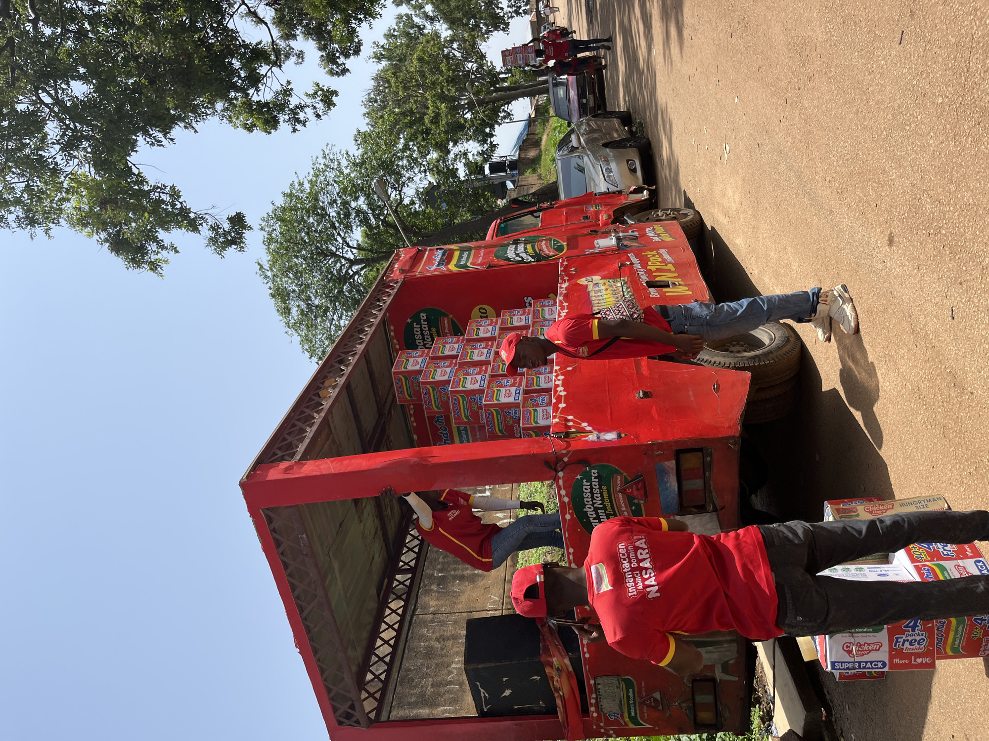 In Rafin Par, Nigeria, a red truck is parked on a dusty road, its bed filled with stacks of Indomie instant noodle boxes. Two men in red shirts with yellow trim, one wearing a red cap, are actively unloading the goods. The man with the cap is facing away from the camera, holding a phone to his ear. The other man, also wearing a red shirt and cap, is inside the truck bed, helping to offload the boxes. Another man in a red shirt and blue jeans is standing near the truck, holding a stack of noodle boxes. Further down the road, other people are visible, some carrying boxes and walking, and a few cars are parked. The scene is illuminated by bright daylight under a clear sky, with trees lining the side of the road. The truck has branding that includes "Indomie" and other text in what appears to be a local language.