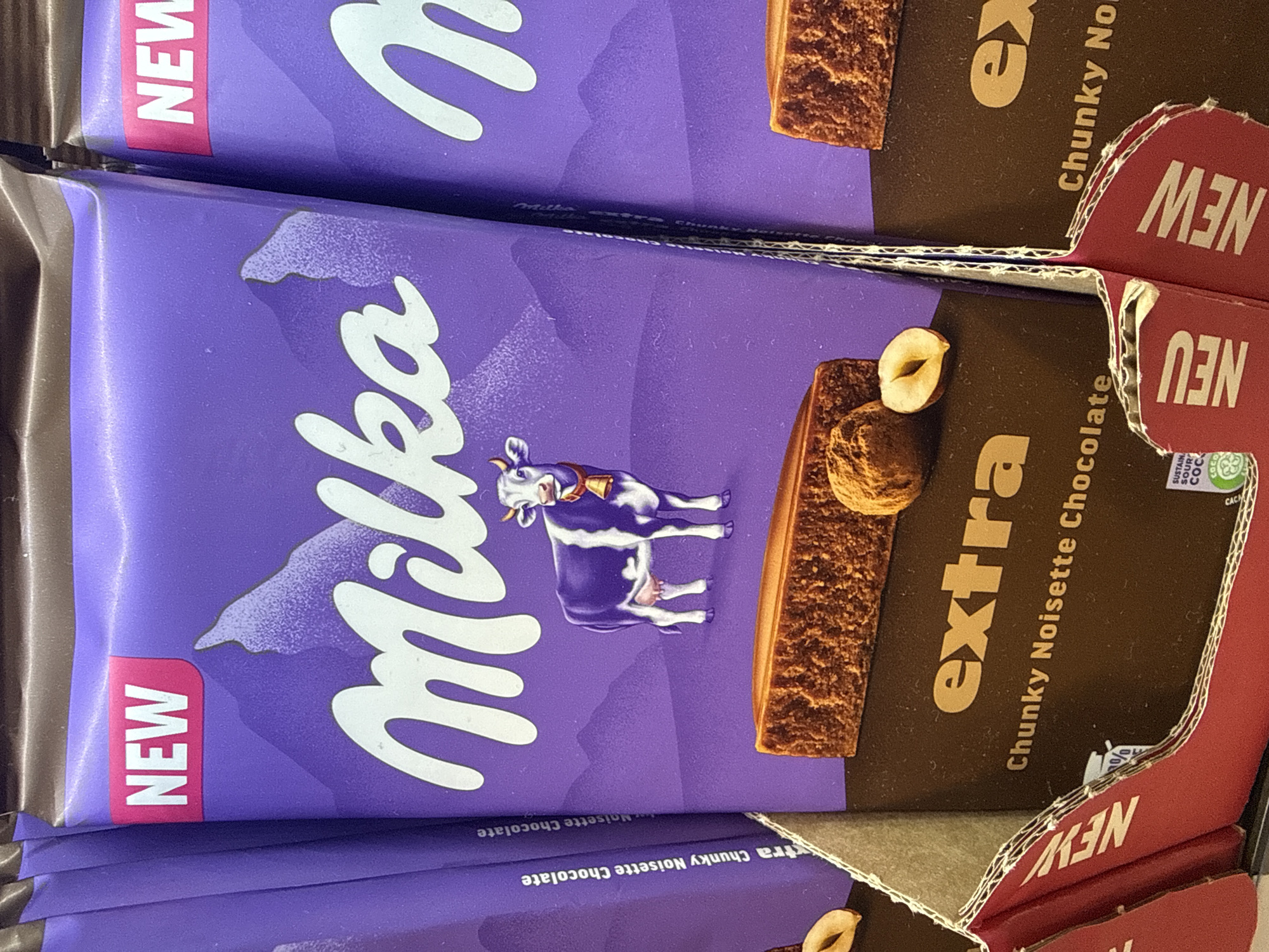 The image shows a close-up shot of several Milka chocolate bars, specifically the "extra Chunky Noisette Chocolate" variety. The packaging is primarily purple with a depiction of the Milka cow. The word "NEW" is prominently displayed on the packaging in red. A hazelnut and a piece of the chocolate bar are shown on the front of the packaging. The bars are stacked in a display case with a cardboard divider, also labeled "NEW" in red. The chocolate bars were photographed in Bulgaria; however, the location, city, and place were not specified.