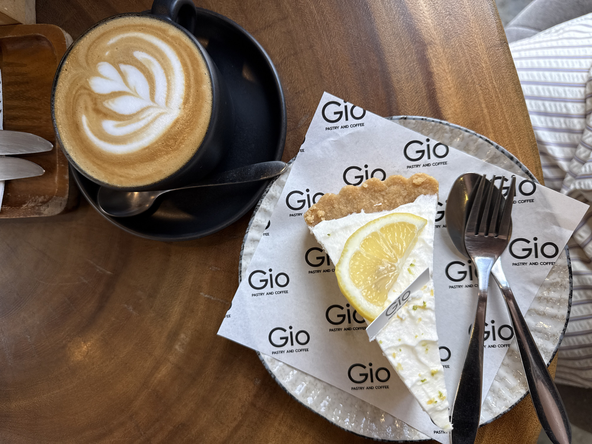 A slice of lemon tart and a cup of latte sit on a wooden table. The tart, on a speckled plate, is covered with a paper wrapper featuring the "Gio Pastry and Coffee" logo. A slice of lemon adorns the tart. A spoon rests on the saucer of the black coffee cup, which contains a latte with latte art. A fork and spoon are placed on the plate.