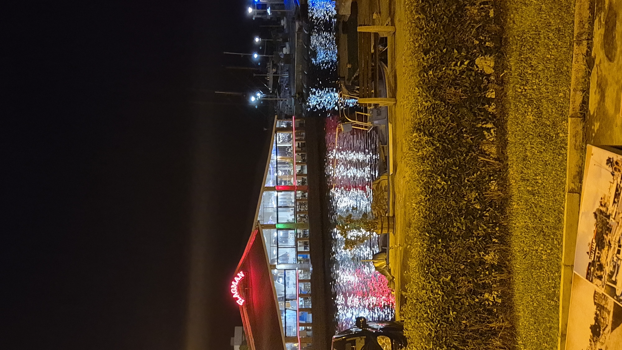 The image depicts a nighttime scene in Nesebar, Bulgaria, featuring a waterfront restaurant named "FLAGMAN" illuminated by red neon signage. The restaurant, with its glass facade, shows tables and chairs inside, suggesting it is open for business. The reflections of colorful lights from the restaurant and other sources shimmer on the dark water of the harbor. In the foreground, a black car is partially visible on the left, and a small boat with "MOPO" written on it is docked on the right. A wooden bench and a potted plant are also present along the waterfront. Behind the waterfront area, a dense hedge and a strip of grass are visible. The sky above is dark, with a few distant lights suggesting the presence of other buildings or structures. The overall impression is that of a lively evening ambiance at a coastal dining establishment.
