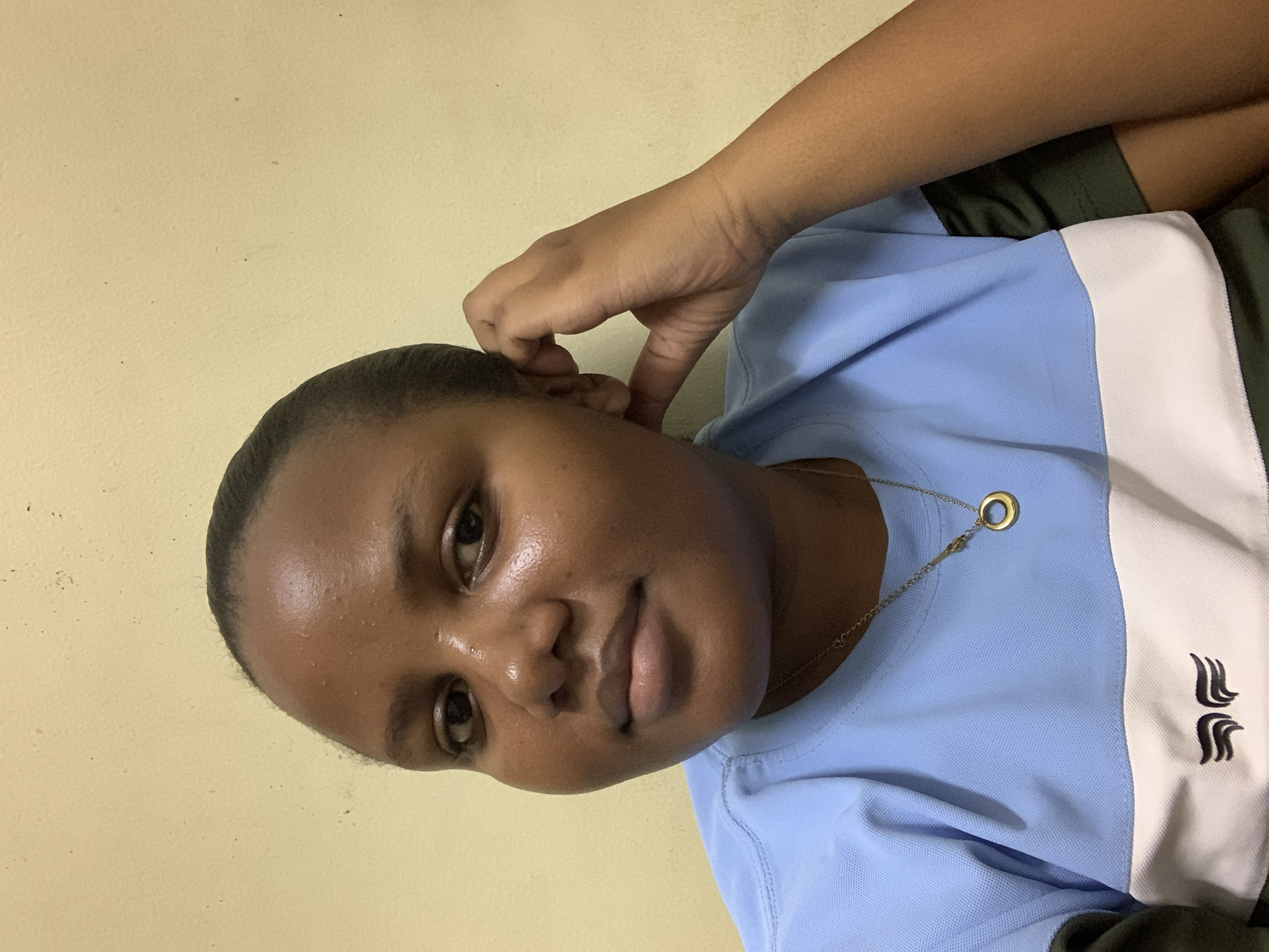 A medium shot features a dark-skinned woman with bare hair. She wears a pale blue polo shirt with white and olive green accents on the right side. A gold chain with a circular pendant rests on the front of her shirt. Her right hand is raised, with fingers near her ear. The background consists of a plain, light beige wall. The photo seems to be taken indoors under soft lighting. There is no information given in this image about the location in Dar es-Salaam, Tanzania.