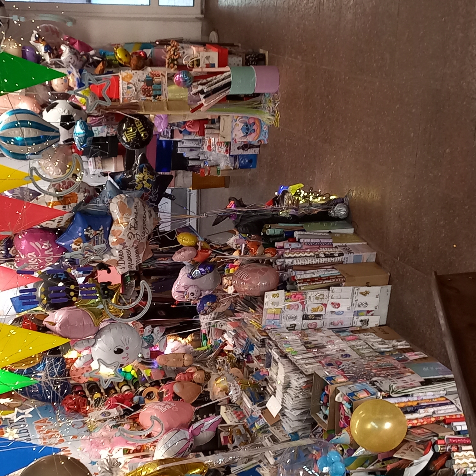 The image shows a stall selling a variety of party supplies, primarily balloons, in a dimly lit indoor market or shop. The stall is densely packed with colorful balloons of different shapes and sizes, including foil balloons shaped like animals, numbers, and abstract designs. There are also several strings of fairy lights illuminating the display. In the background, other market stalls are visible, with shelves stacked with various goods. The floor is made of a reddish-brown material, possibly tile or concrete, and appears somewhat worn. The overall impression is one of a busy, festive, and slightly cluttered retail environment. There are no people clearly visible in the frame. The location is described as Kropyvnytskyi, Ukraine.