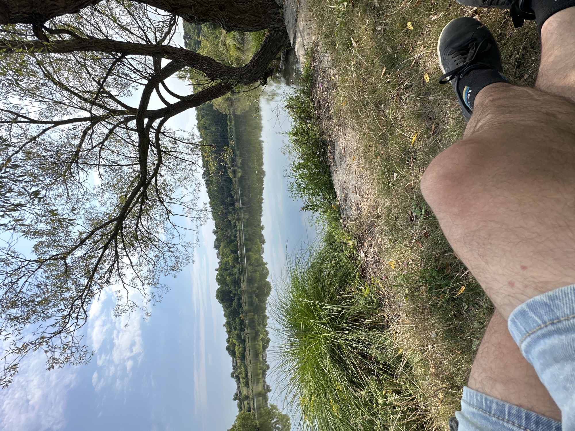 A person's legs are visible in the foreground, clad in denim shorts and black athletic shoes with black socks. They are seated on a grassy bank overlooking a calm body of water. The water reflects the surrounding trees and the blue sky with scattered clouds. In the background, a treeline with scattered houses lines the opposite shore. The scene appears to be a natural landscape, possibly a park or recreational area, in or near Makiv, Ukraine.