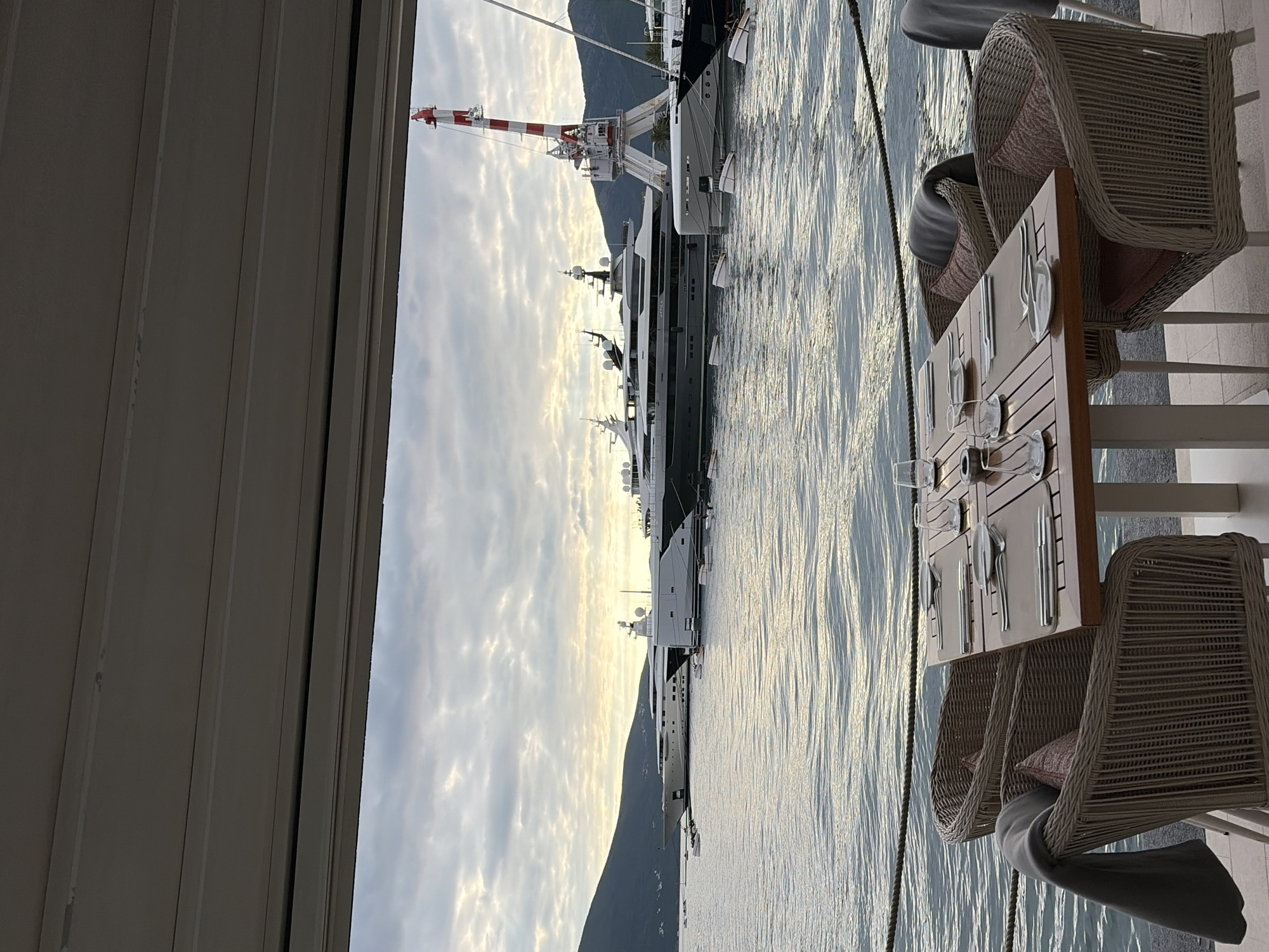 The image depicts an outdoor dining area in Tivat, Montenegro, overlooking a marina. The scene is observed from beneath a covered area, possibly a restaurant patio. Several yachts of various sizes are docked in the water. A large industrial crane is located near the yachts. The water is calm with reflections of the sky. A dining table is set for a meal, with cutlery and glassware arranged on it. Two wicker chairs with cushions are positioned at the table. A mountain rises in the background. The sky is overcast with clouds.