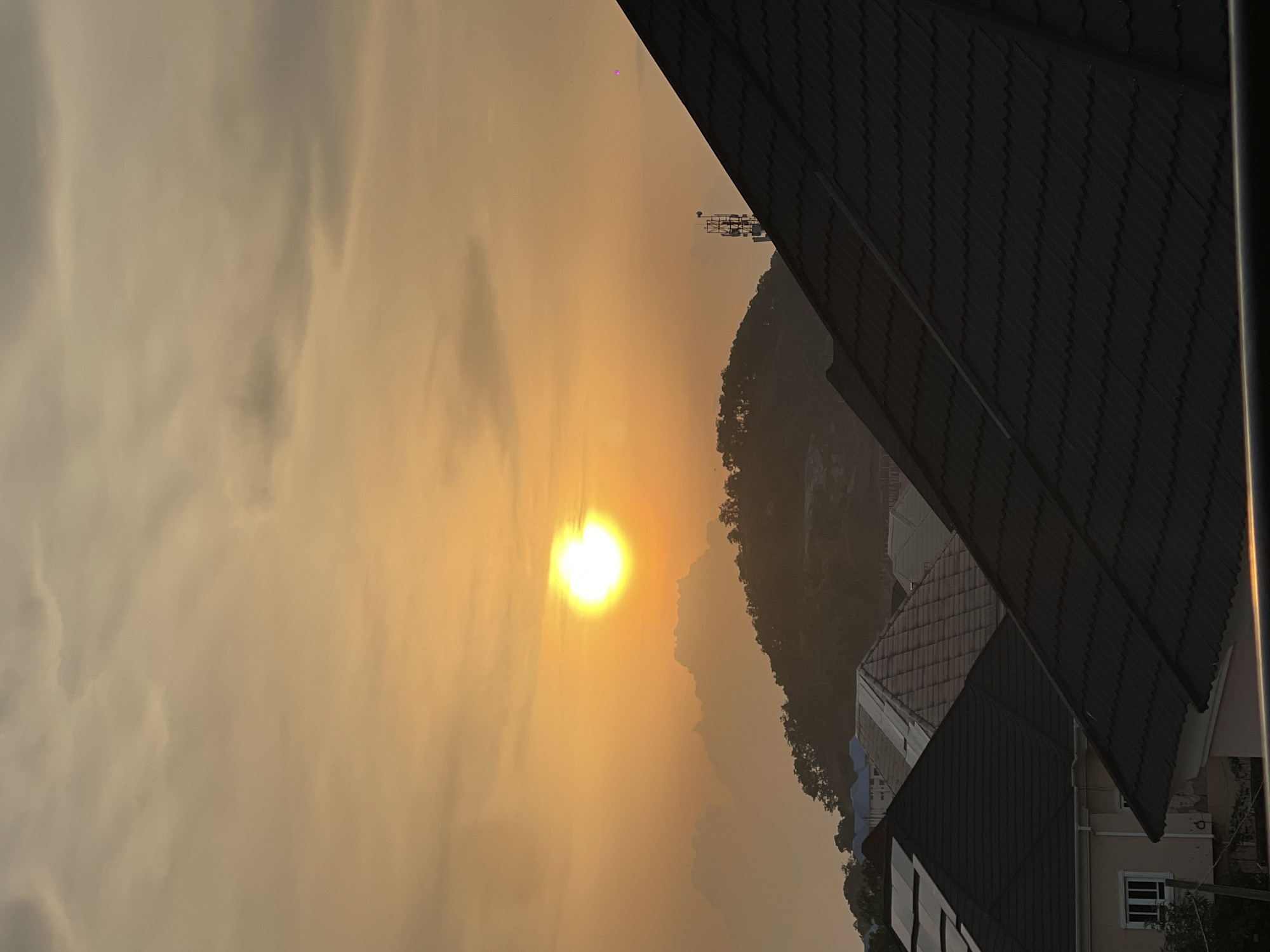 The image depicts a sunset in Abuja, Nigeria. The sun, a bright, radiant orb, is positioned in the upper center of the frame, casting a warm, golden-orange glow across the sky and clouds. The sky is a blend of soft oranges and muted grays, indicative of a hazy or dusty atmosphere common in the region.
In the midground, a tree-covered hill or small mountain rises, silhouetted against the vibrant sky. Its contours are softened by the haze. On the slopes of the hill, a telecommunications tower is visible, its metallic structure standing out against the natural landscape.
In the foreground, the tiled roofs of several houses are prominent. The roofs are dark, with a corrugated texture, and are arranged at varying angles, creating a sense of depth and urban sprawl. Some houses have visible windows and walls, with one house in the lower left corner showing a light-colored facade and a white-framed window. The overall impression is of a residential area at the edge of a more natural, elevated landscape during the golden hour. No people are visible in the image.
