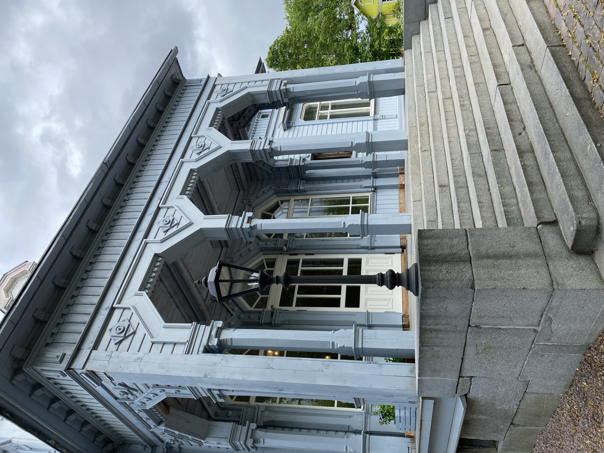 The image shows a light blue wooden building with intricate decorative elements, including carved trim and arched doorways. A wide, stone staircase leads up to the entrance, flanked by large stone blocks on the left. A black lamppost stands prominently on the staircase in front of the entrance. The building has large windows and a white double door. To the right of the building, green trees are visible, and a yellow building can be seen in the distance. The sky is overcast. The building is likely the Vyborg Architectural and Ethnographic Museum-Reserve located in Sortavala, Russia, which was once part of Finland.