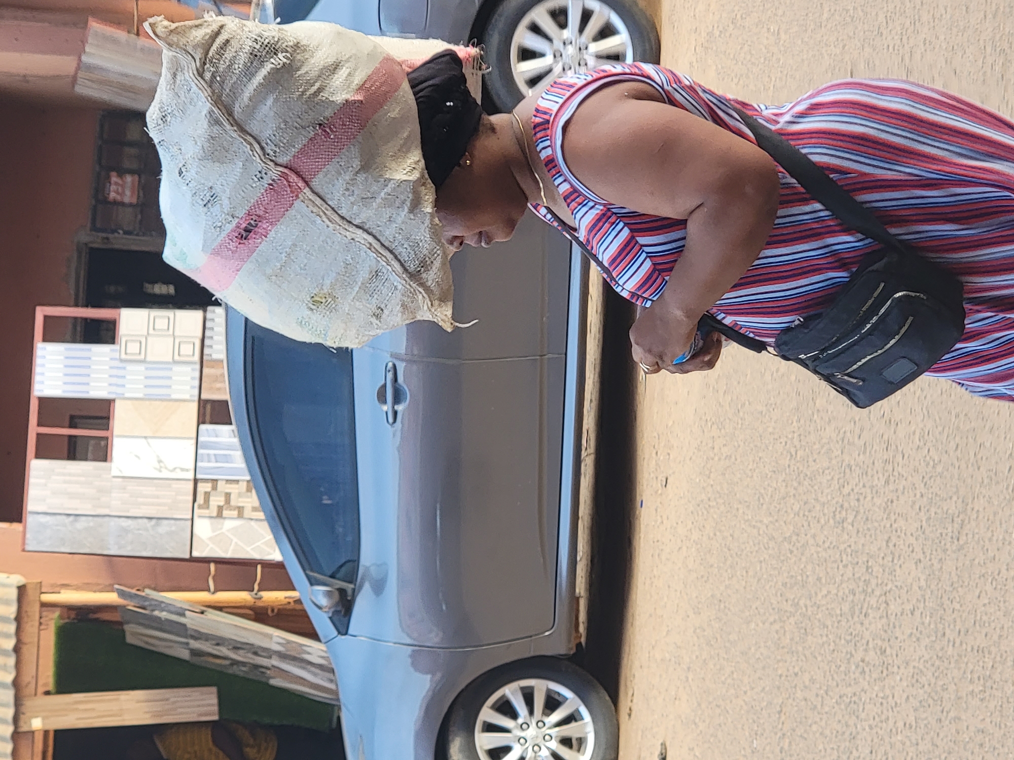 In Maiburiji, Nigeria, a woman with a striped sleeveless dress is carrying a large sack on her head, along a road where a gray car is parked. The woman is wearing a black over-the-shoulder purse. Behind the car, there's a wall display of square tiles.