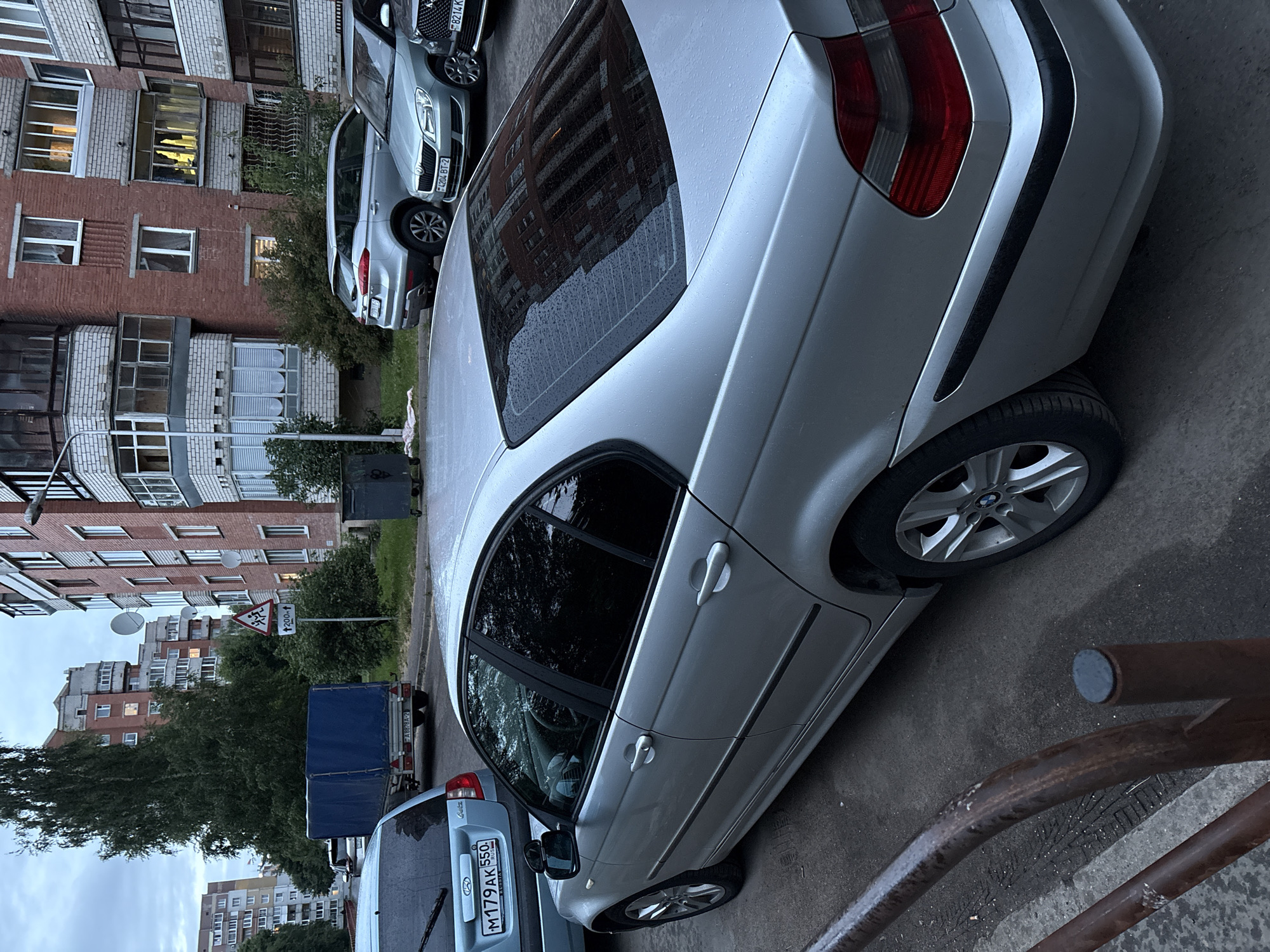 The image depicts a street scene in Vitsebsk, Belarus, likely a residential courtyard. The foreground is dominated by the side and rear of a silver BMW sedan. The car's doors, windows, and wheels are clearly visible. To the left, partially obscured, is the rear of a blue hatchback with a license plate ending in "AK 550". Further back, a blue trailer with a tarp is parked. The background features a multi-story brick apartment building with numerous windows and balconies. Trees and other greenery are interspersed between the buildings. A traffic sign indicating children crossing is visible. The sky is overcast, suggesting it might be evening or a cloudy day, and there are visible water droplets on the cars and surfaces, indicating recent rain. A rusted metal fence runs along the left edge of the frame. The overall scene is one of a parked car in an urban residential area.