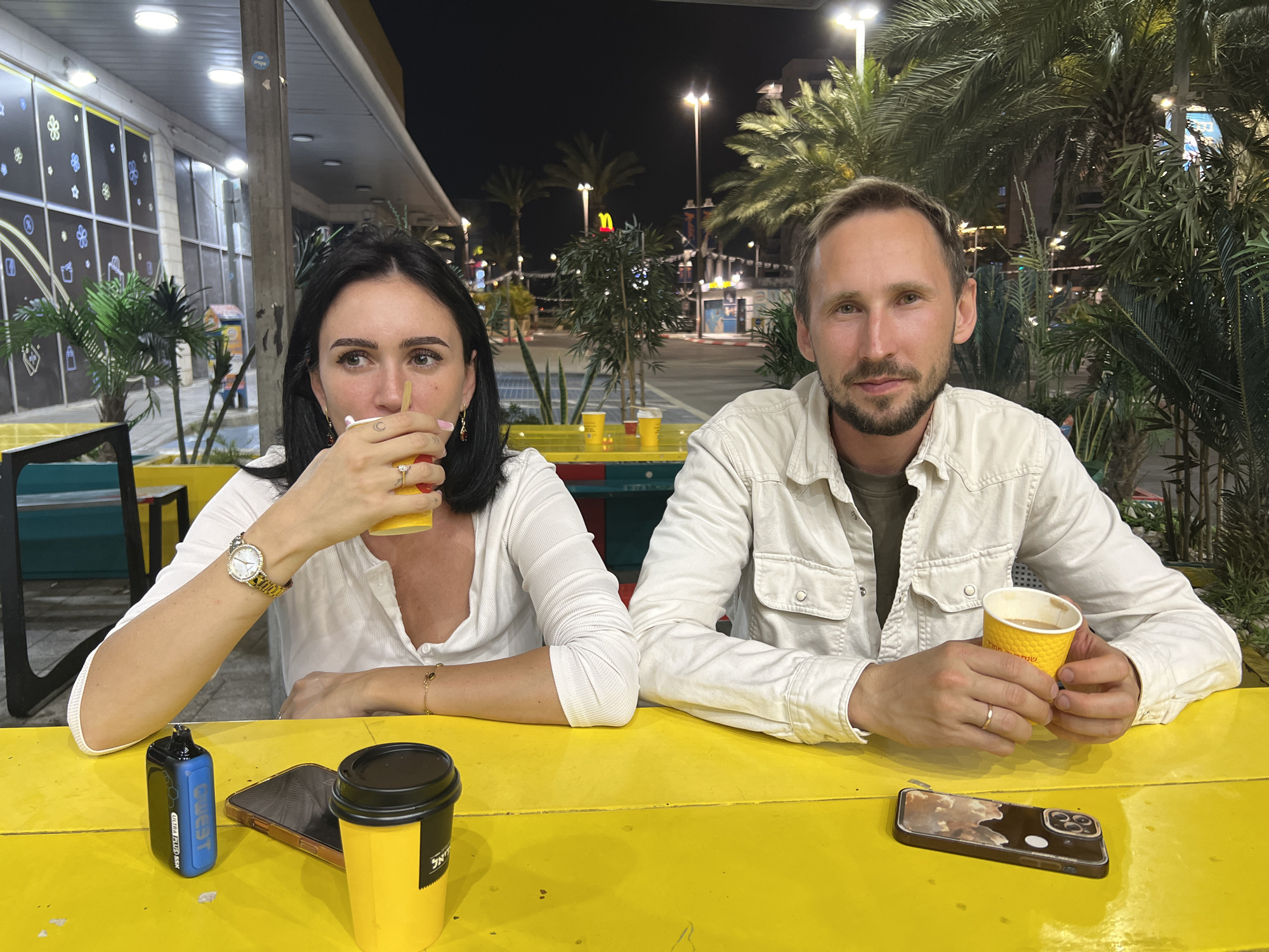 A couple is seated at an outdoor table in Kiryat Motzkin, Israel. The woman on the left has dark hair and wears a white top, a gold watch, and jewelry. She holds a yellow cup to her lips. To her left are a blue vape device, a smartphone, and a yellow cup with a black lid. The man on the right has light brown hair, a beard, and wears a white button-down shirt. He holds a yellow cup in his hands. A smartphone is placed on the table in front of him. The table is painted yellow. The background shows a street scene at night with lights, palm trees, and buildings. A McDonald's restaurant is visible in the background. The building behind the woman has windows with decorative designs.