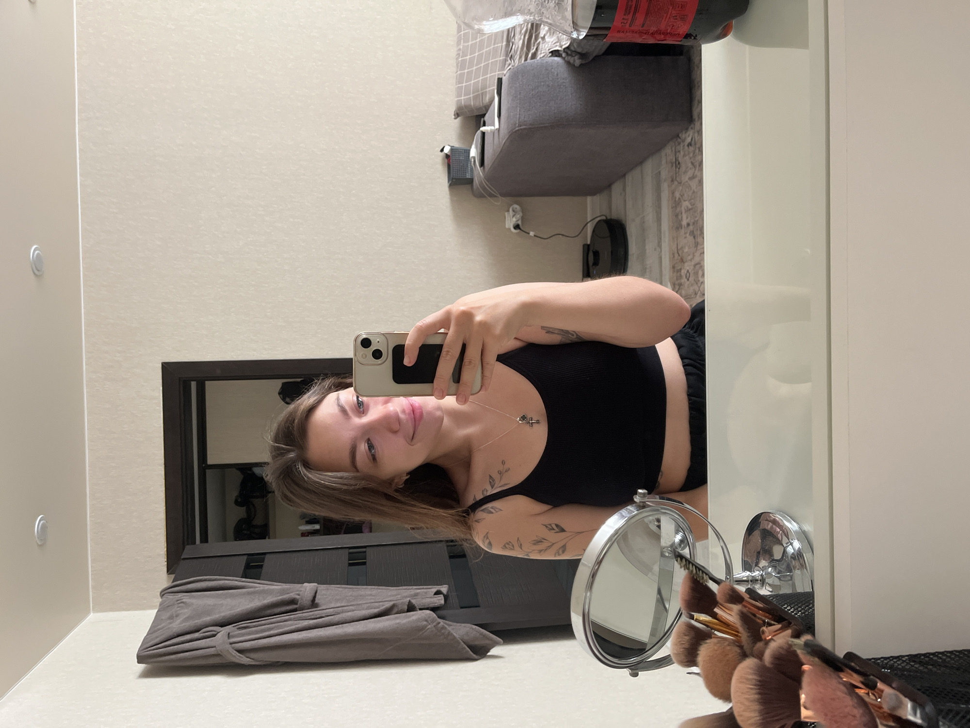 The image depicts a young woman with brown hair, wearing a black tank top and black shorts, taking a mirror selfie. She has tattoos on her left arm and shoulder, and is wearing a necklace with a cross pendant. In the foreground, a makeup brush organizer sits on a white vanity. Behind her, a closet door is partially open, revealing clothes inside. A grey bathrobe hangs on the wall to the left. In the background, a grey couch with patterned cushions is visible, along with a robotic vacuum cleaner on the floor. A large bottle of what appears to be Coca-Cola is positioned on the right side of the frame. The overall environment suggests a residential indoor setting. The scene is set in an apartment in the city of Chaikovsky, Russia.