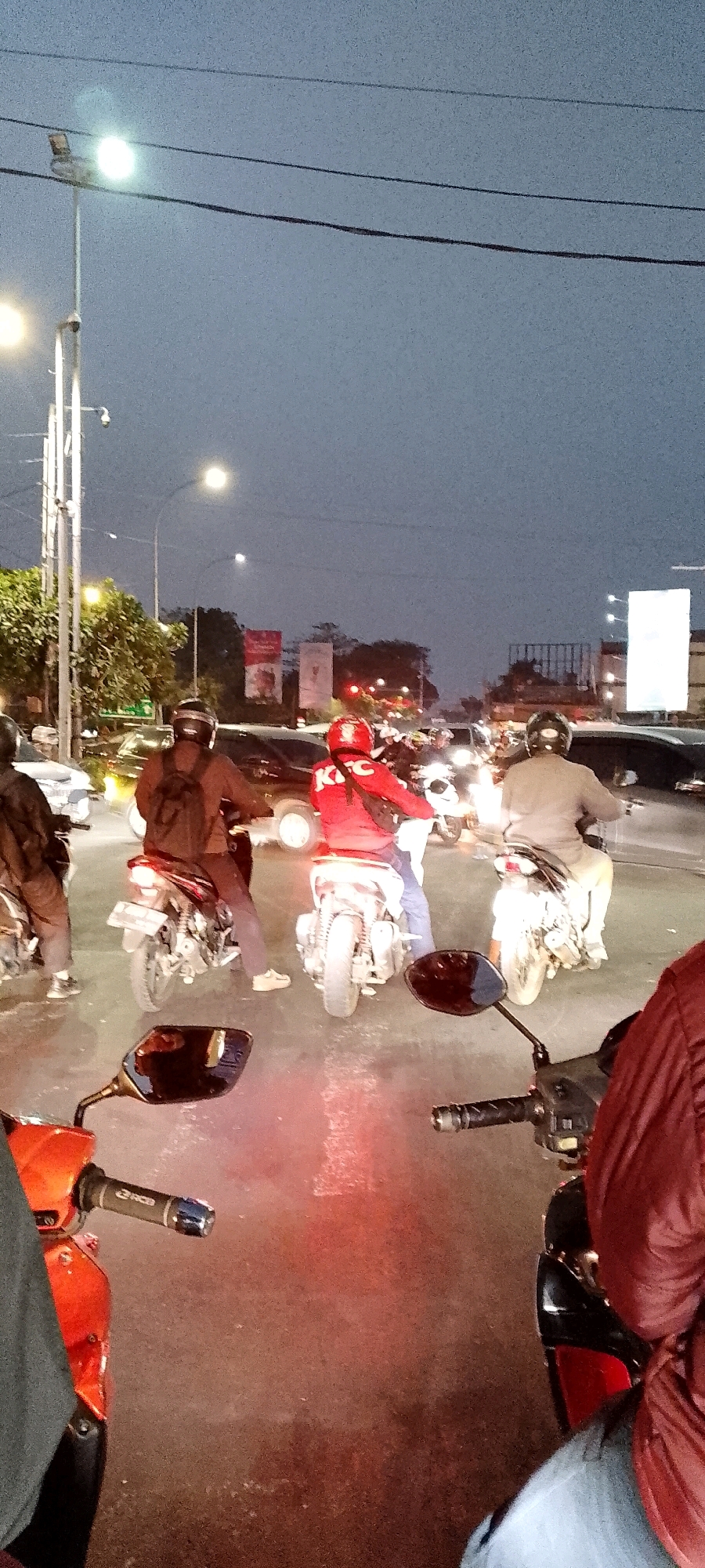 The image captures a scene at dusk in Depok, Indonesia, likely on a street with traffic. Several motorcycles are present, some with riders. One rider in the foreground is wearing a red jacket with white "KFC" lettering. Another rider in the center wears a brown jacket and a black helmet, and is on a motorcycle with a license plate. The environment includes streetlights, overhead wires, and trees. The road appears wet, possibly indicating recent rain. The lighting suggests it's either early morning or late evening. In the background, there are buildings and billboards, typical of an urban setting. The perspective suggests the photo was taken from another motorcycle in traffic.