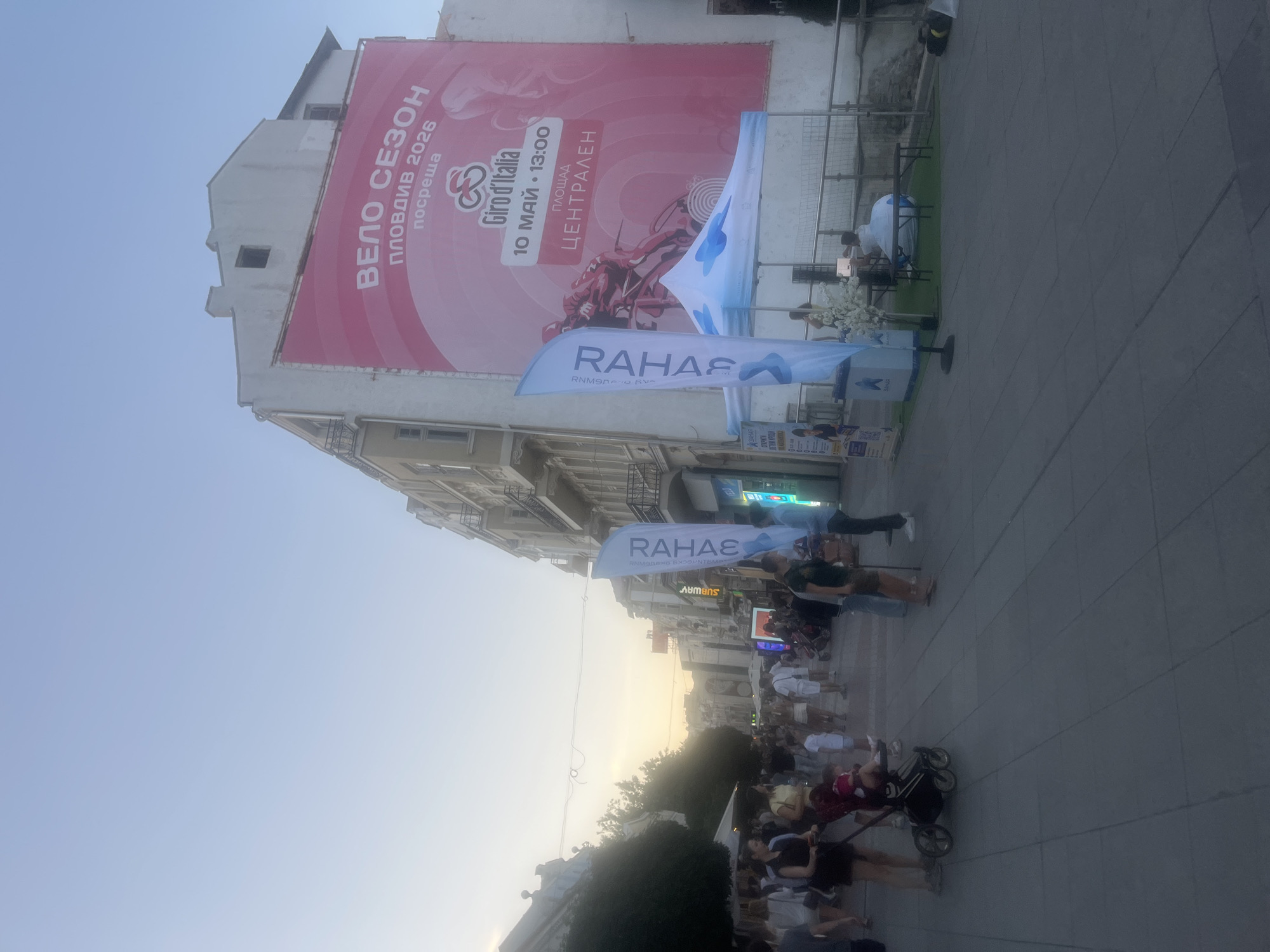 In an outdoor urban setting, a wide street is lined with multi-story buildings. A large banner on the side of one building advertises "VELO SEZON PLOVDIV 2026" and "Giro d'Italia" with a date and time of "10 MAY 13:00". A crowd of people are walking along the paved street, some pushing strollers. Two blue flags with white text stand near a white tent. In the distance, more buildings and trees are visible under a pale sky. The lighting suggests it is late afternoon or early evening.