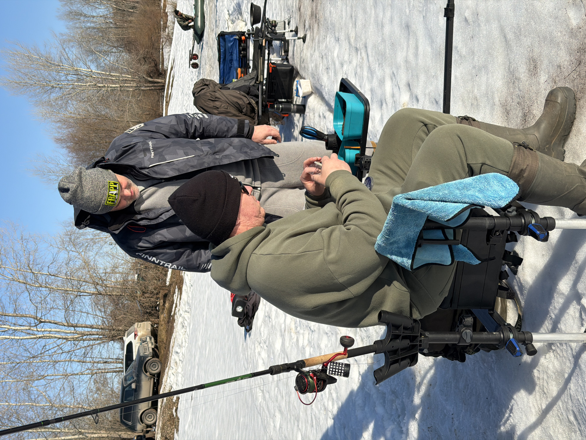 Two men are ice fishing in a snowy area, likely near the city of Chaikovsky, Russia. One man is wearing a grey and black "Finntrail" jacket and a grey "Frapp" beanie. The other man is wearing a black beanie and an olive green hooded jacket. They are both seated on fishing chairs, with fishing rods and gear visible around them. The ground is covered in snow, and bare trees are visible in the background. In the distance, a vehicle is parked near the edge of the snowy area.