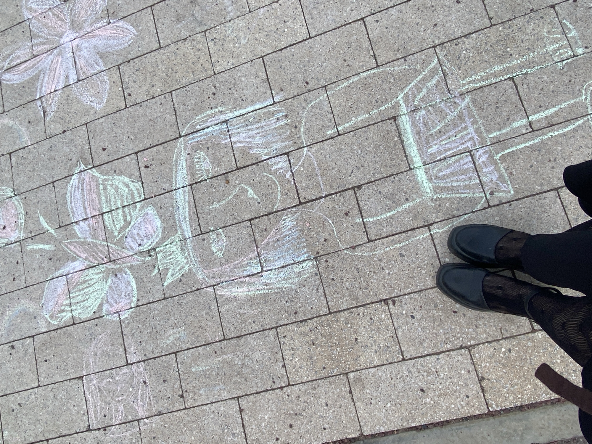 The image shows a paved sidewalk with chalk drawings, presumably in the city of Chaikovsky, Russia. The drawings include a flower, a face, and what appears to be a building or structure. A pair of legs wearing black tights with a pattern and black shoes are visible on the right side of the image, suggesting a person standing near the drawings. The environment consists of grey concrete pavement. No interaction is explicitly visible, but the chalk drawings imply someone drew on the pavement. No other people or actions are visible in the frame.