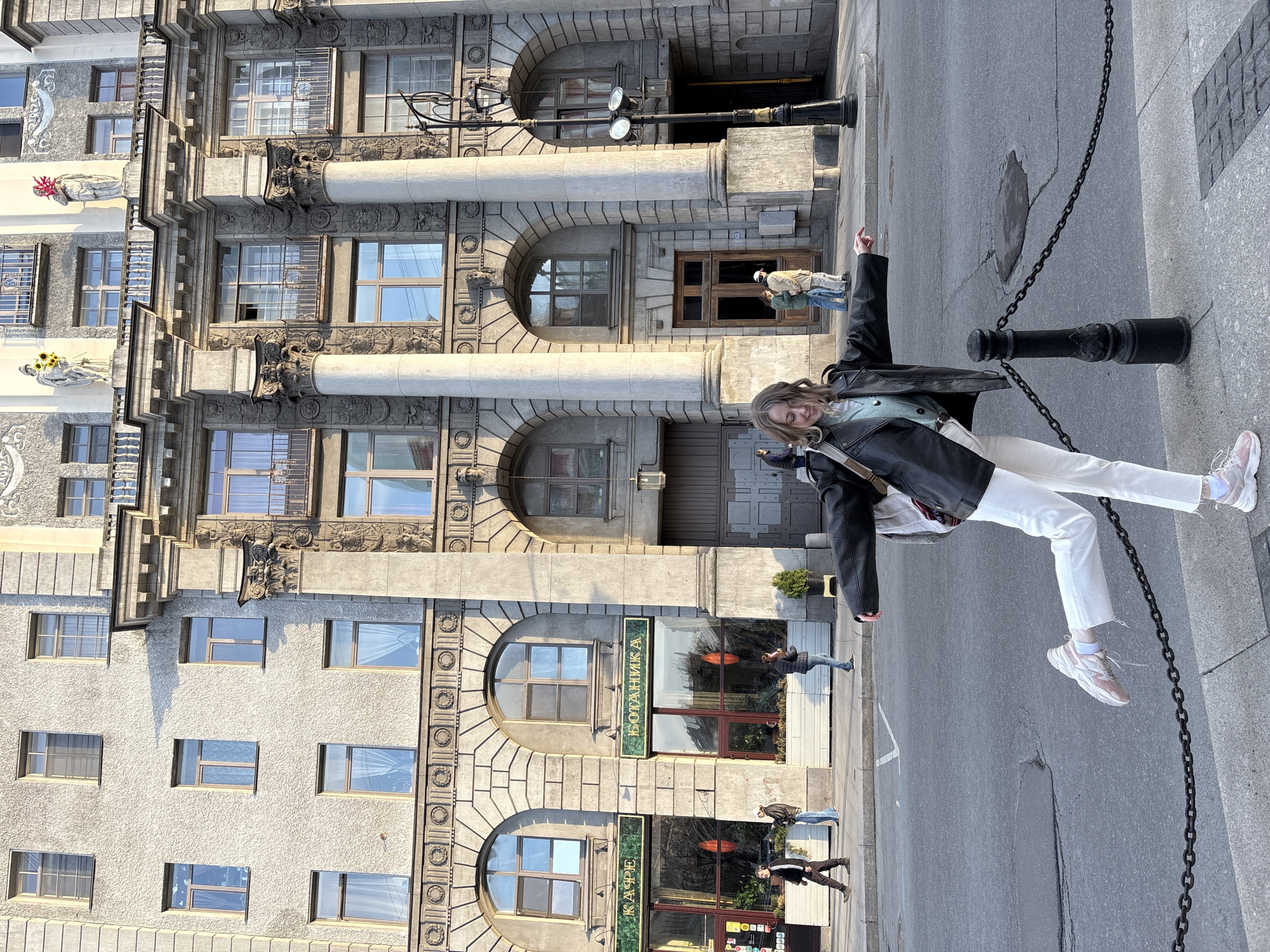 The image is of a woman standing in front of a building in Saint Petersburg, Russia. The building is a light grey color with ornate architectural details. The woman is wearing a black leather jacket, white pants, and pink sneakers. She is leaning against a black metal post with a chain attached to it. The background includes pedestrians and the street. The building appears to house a cafe.