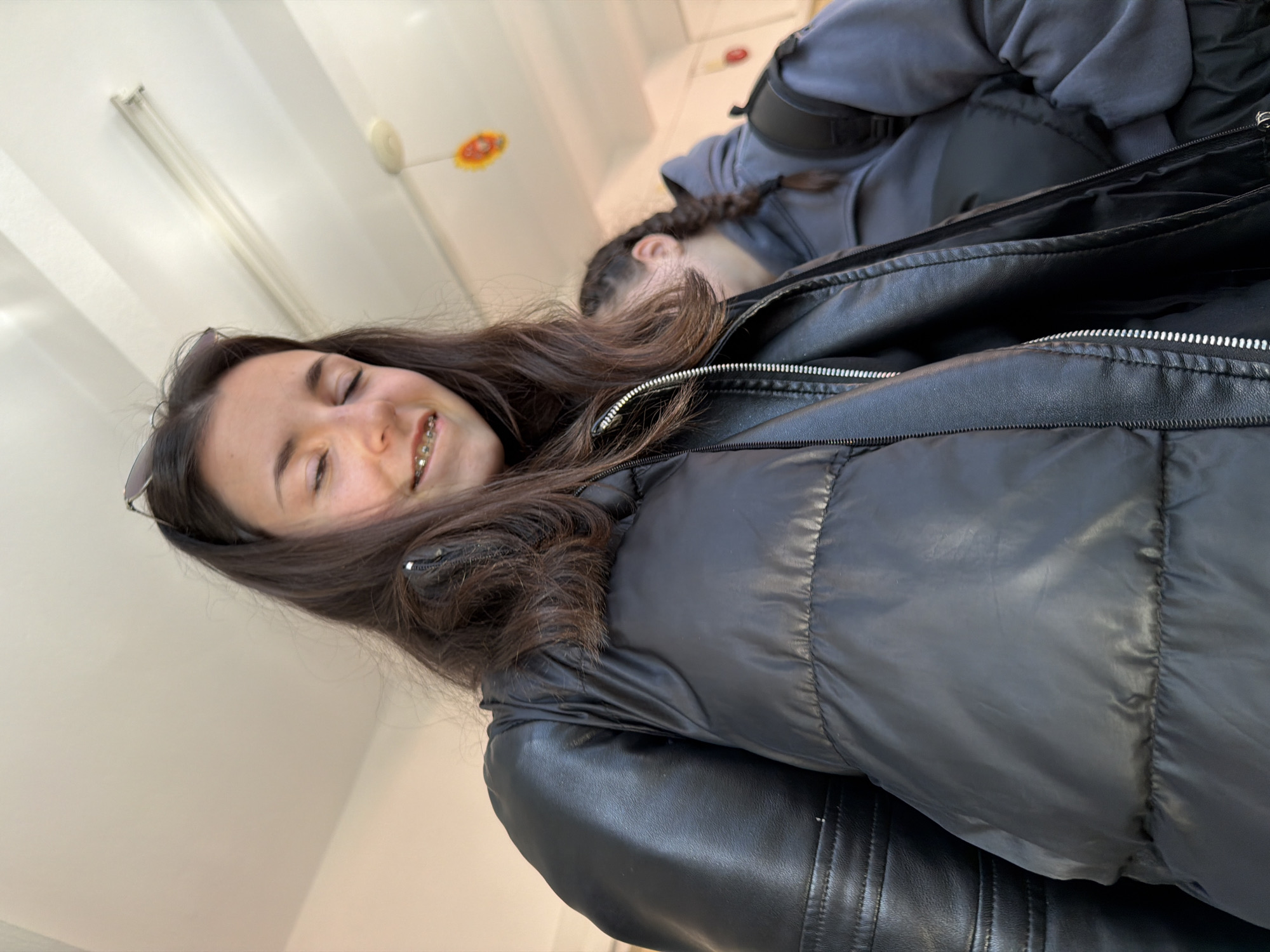 The image depicts two young women indoors, possibly in Panagyurishte, Bulgaria. One woman is prominently featured in the foreground, smiling with braces visible. She is wearing a black leather jacket with a puffy black vest layered over it. A pair of sunglasses is perched on her head. Behind her and slightly out of focus is another woman with her hair in a braid, wearing a grey hooded sweatshirt and a backpack. The background shows a white wall with fluorescent lighting fixtures. A decorative flower is visible near the ceiling. The overall scene suggests a casual setting.