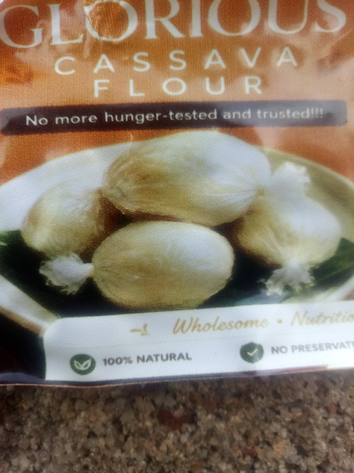 The image is a close-up photograph of packaging for "Glorious Cassava Flour". The packaging features text that reads "GLORIOUS CASSAVA FLOUR" prominently at the top, followed by "No more hunger-tested and trusted!!!". Below this, there is an image of what appears to be several small, wrapped food items, possibly dumplings or parcels, arranged on a plate or in a bowl.
Further down on the packaging, there is a white banner with text and icons. Visible text includes "Wholesome", "Nutrition", "100% NATURAL", and "NO PRESERVAT". There is also a green leaf-like icon next to "100% NATURAL" and a green checkmark icon next to "NO PRESERVAT".
The background of the packaging appears to be an orangey-brown color with some texture. The overall impression is of a product that is being marketed as healthy, natural, and reliable. The image quality is somewhat blurry, particularly in the lower portions. The overall scene is a product shot focused on the packaging and its advertised benefits. There are no people visible in the image. The lighting seems to be even, suggesting it might be taken indoors with artificial lighting or in an overcast outdoor environment. The texture of what appears to be gravel or sand is visible at the very bottom of the image, suggesting the packaging might be resting on such a surface.