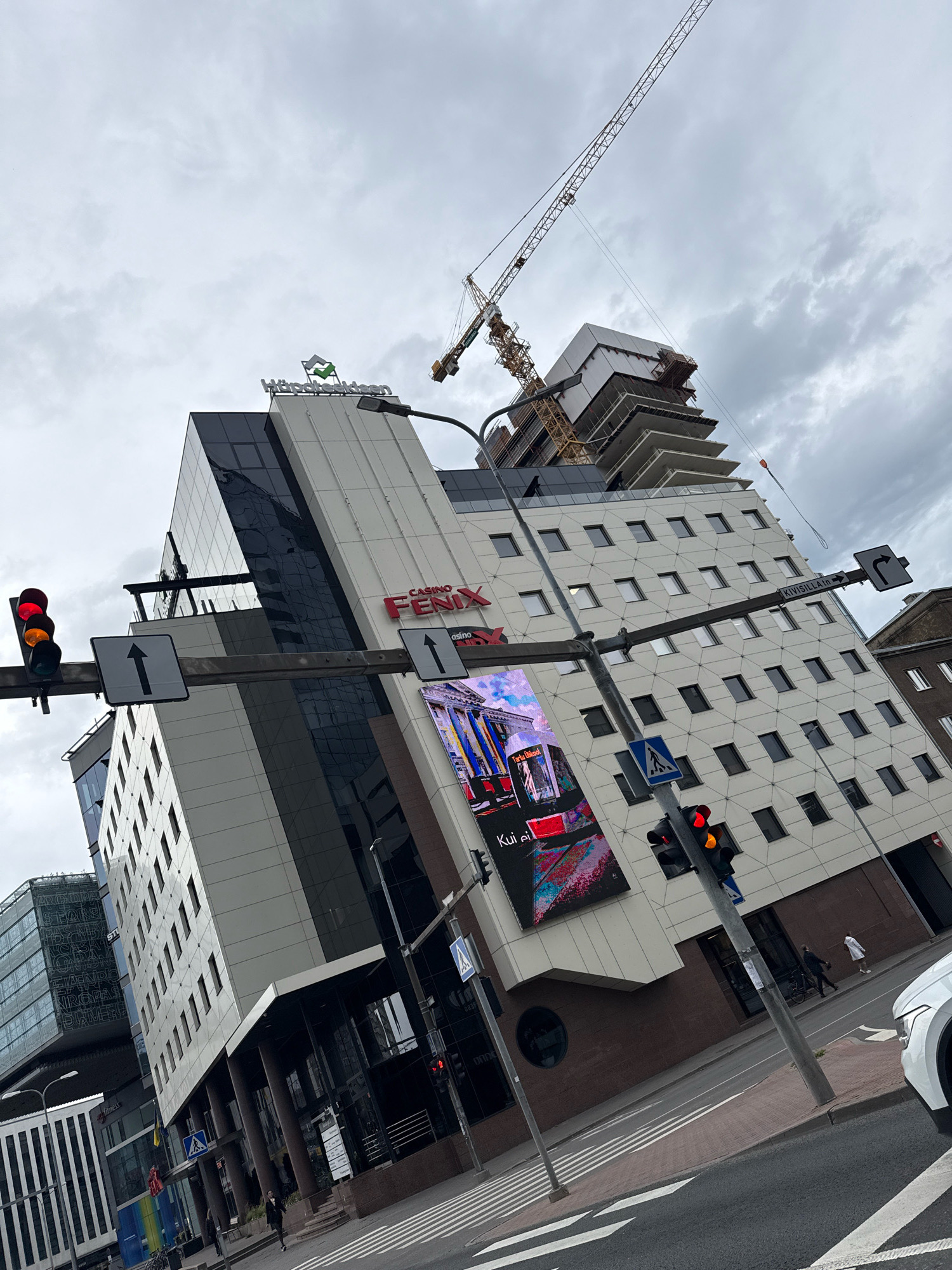 In Tallinn, Estonia, a tilted urban scene unfolds under an overcast sky, dominated by a modern office building under construction. A large yellow crane, a prominent feature of the construction site, looms in the background. The foreground is occupied by a traffic signal displaying a red and amber light, indicating a caution or stop. Adjacent to the traffic signal are two directional signs pointing upwards and to the right. The building itself is a mix of glass and white facade, with the "Casino Fenix" logo prominently displayed. A large digital billboard on the building showcases a colorful image. The street level shows a crosswalk with striped markings, and a few pedestrians are visible, including two individuals walking near the building entrance and another further down the street. A white car is partially visible on the right side of the frame. The architectural style suggests a contemporary urban environment in Tallinn.