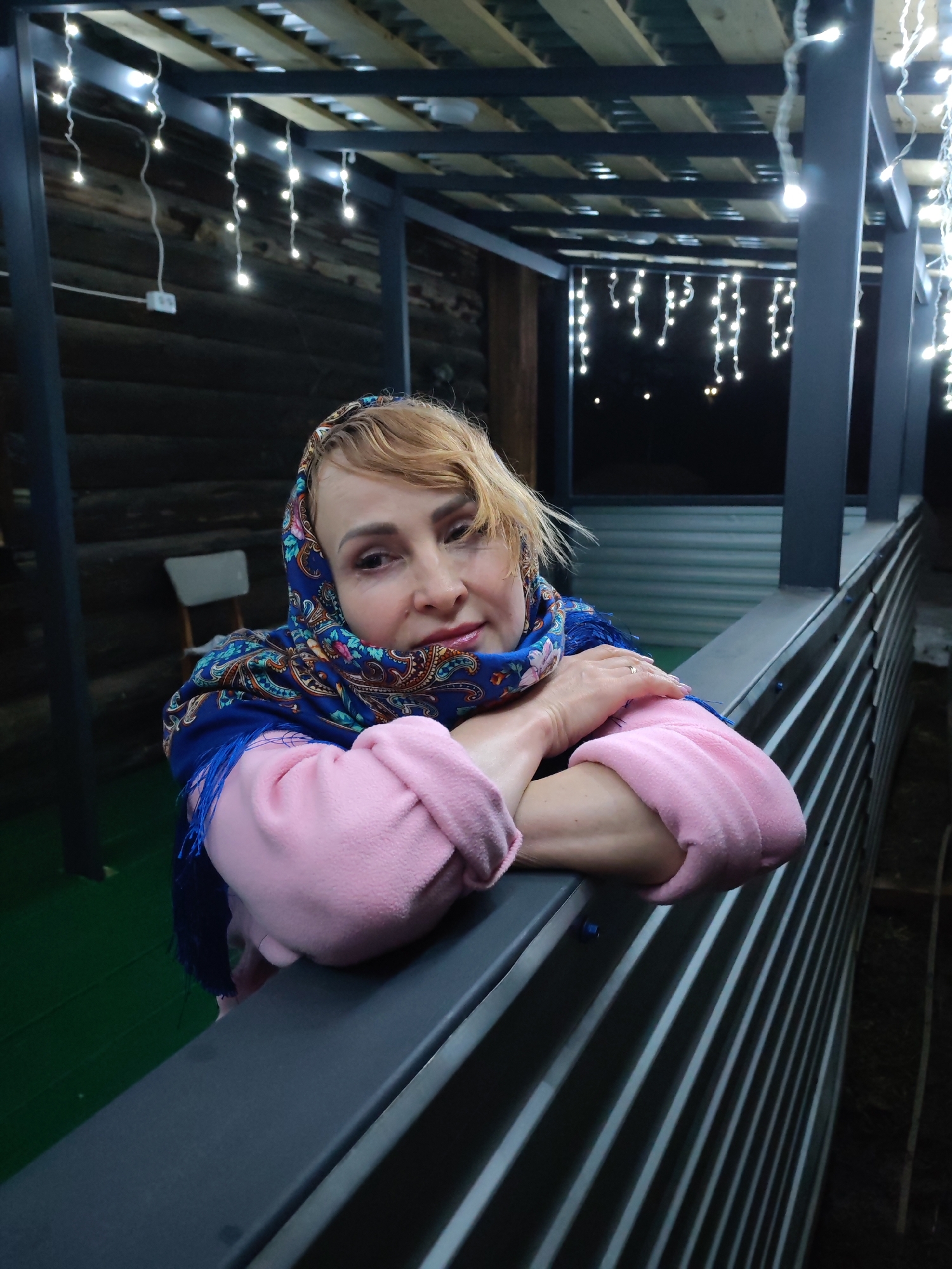A woman wearing a blue headscarf with a floral pattern and a pink sweater is leaning on a grey railing. She is outdoors, under a structure adorned with string lights, which suggests an evening setting. The location is implied to be in the city of Chaikovsky, Russia. The background includes a wooden structure and glimpses of the night sky. The woman is looking towards the camera with a slightly pensive expression, her hands resting on the railing.